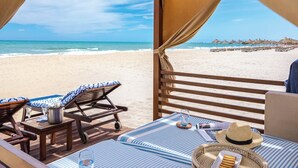 Una habitación frente a la playa, con vista al océano, una silla de lounge, una mesa con un cubo y un vaso, y un sombrero de paja sobre la cama.