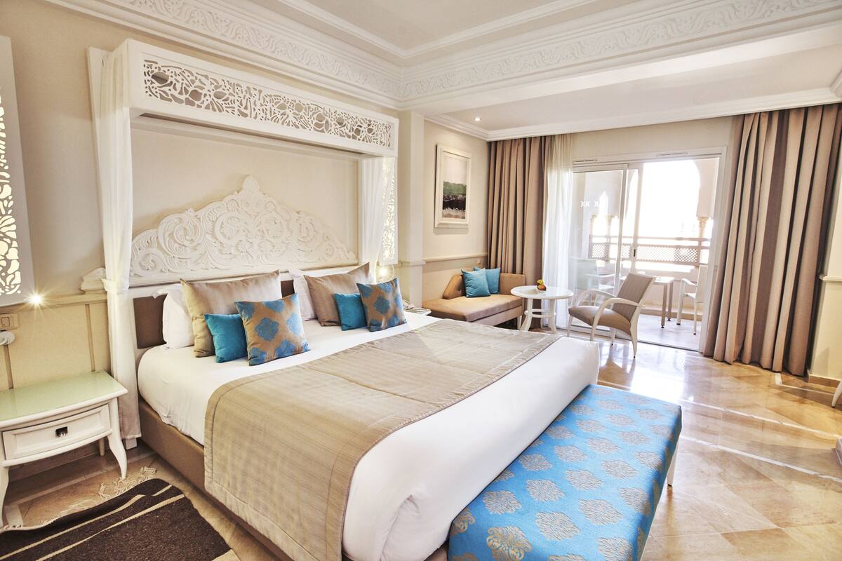 A spacious hotel room with a large bed, a sitting area with a sofa and chairs, a small table, and a balcony with curtains.