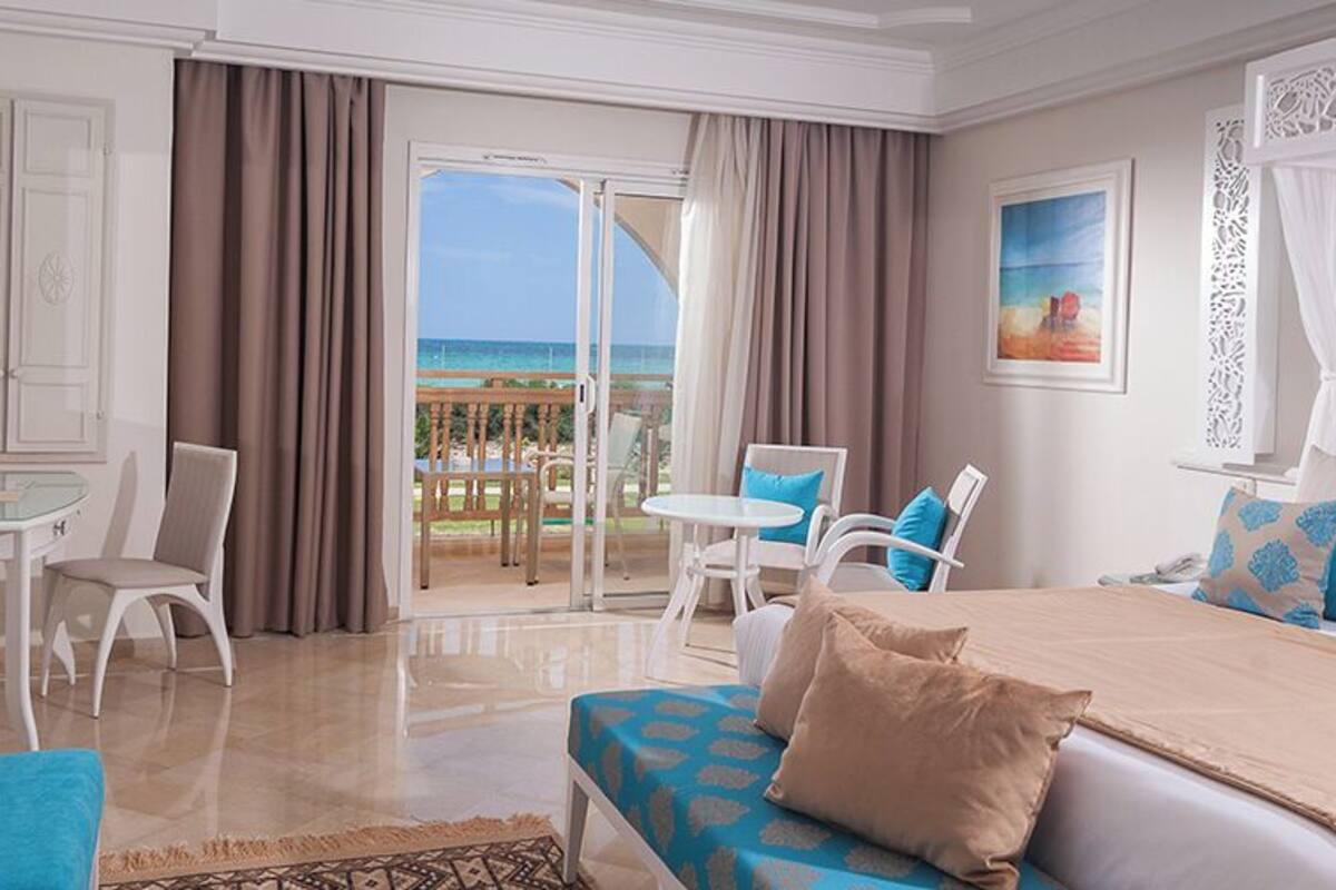 A hotel room with a bed, a desk, a chair, and a balcony with a view of the sea.
