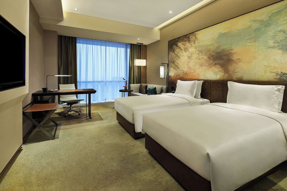 Ein Hotelzimmer mit zwei Betten, einem Schreibtisch, einem Sessel, einem Fernseher und einem großen, abstrakten Gemälde an der Wand.