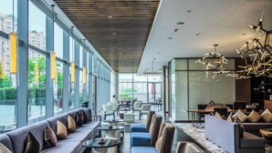 A modern lounge area with a long sofa, armchairs, and tables, large windows offering views of the city, and contemporary lighting fixtures.