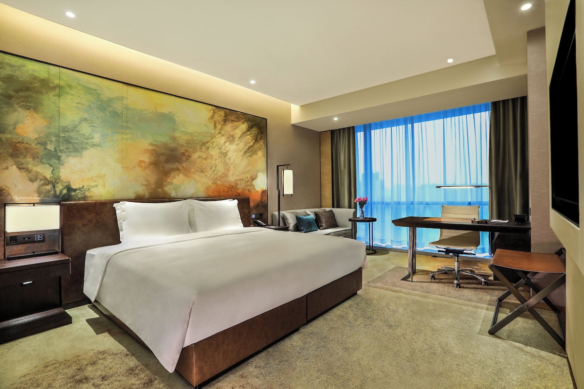 A modern hotel room with a large bed, a desk, and a view of the city through the window.