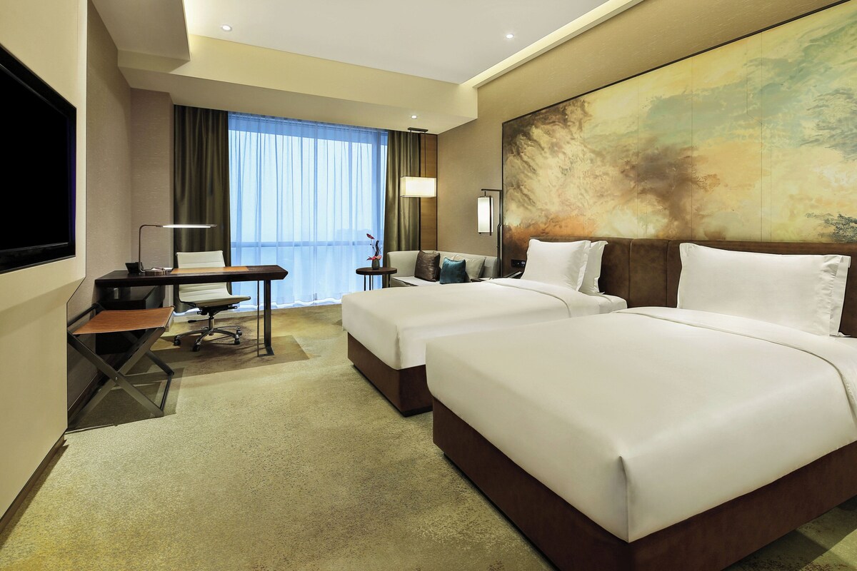 Ein Hotelzimmer mit zwei Betten, einem Schreibtisch mit Stuhl, einem Fernseher und einem großen, abstrakten Gemälde an der Wand.