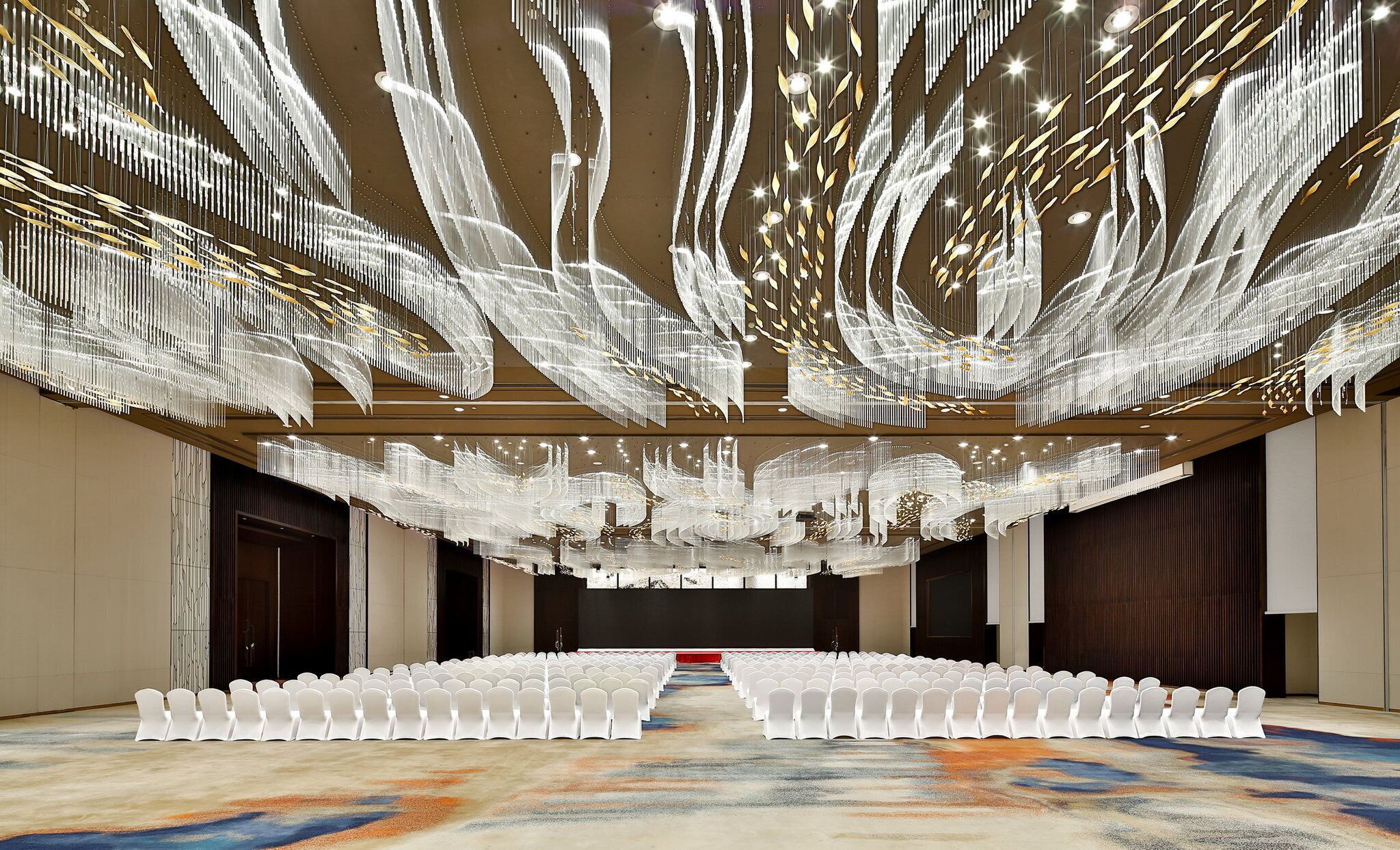 Un amplio salón con filas de sillas blancas, una alfombra estampada y un techo decorado con lámparas colgantes.