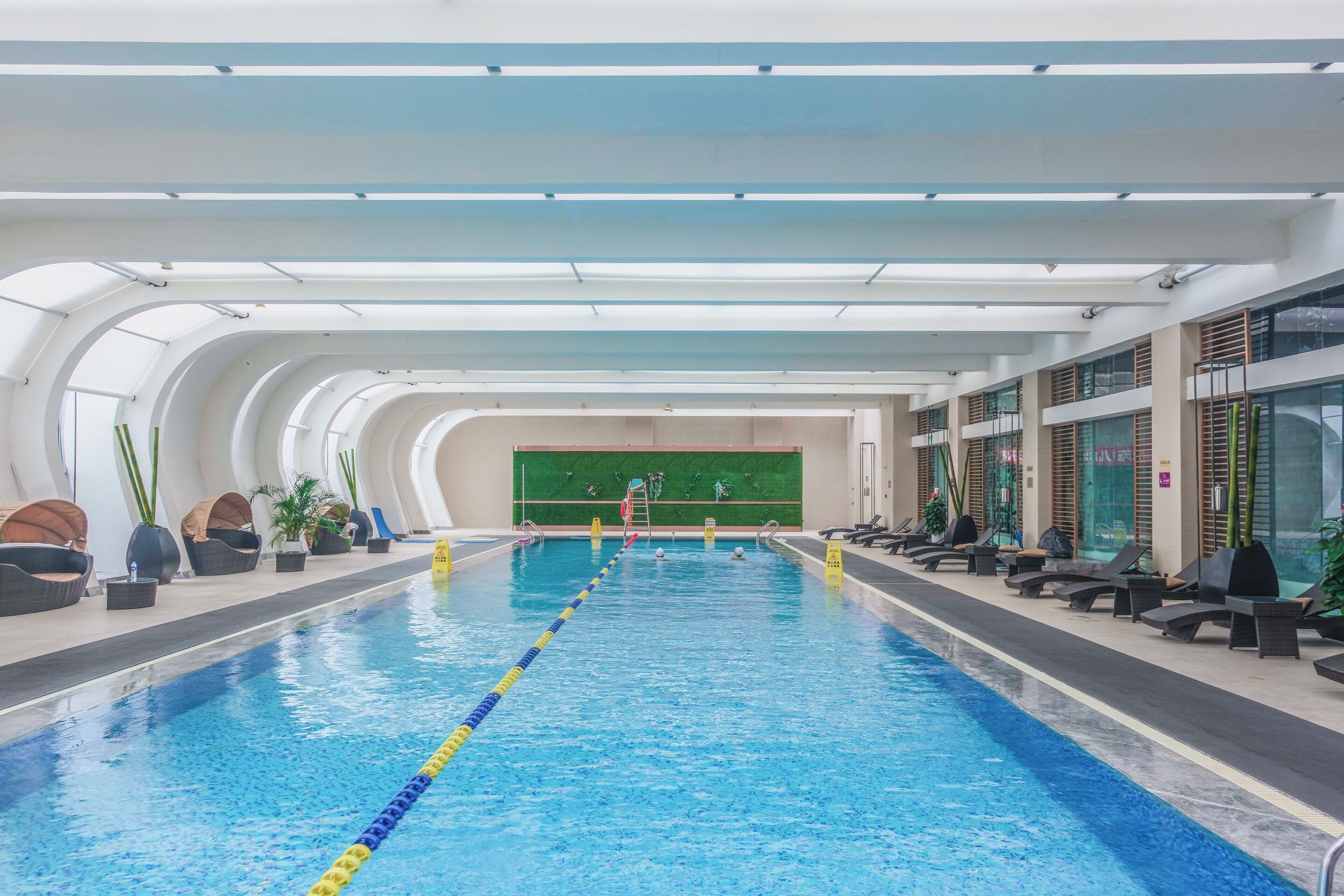 An indoor swimming pool with clear lane dividers, lounge chairs, and potted plants along the sides.