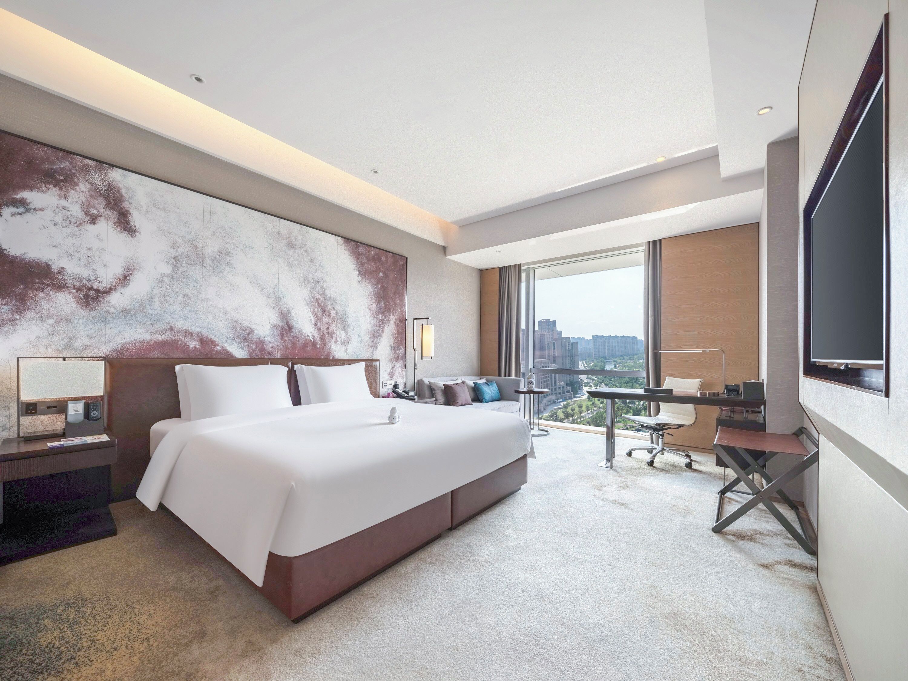 A modern hotel room with a large bed, a desk, a chair, and a large window offering a city view.