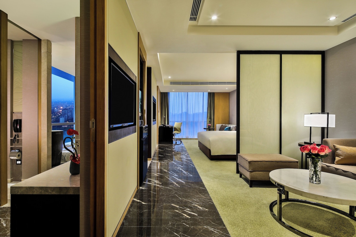 A hotel room with a bed, a desk with a computer, a TV mounted on the wall, and a view of the city at night.