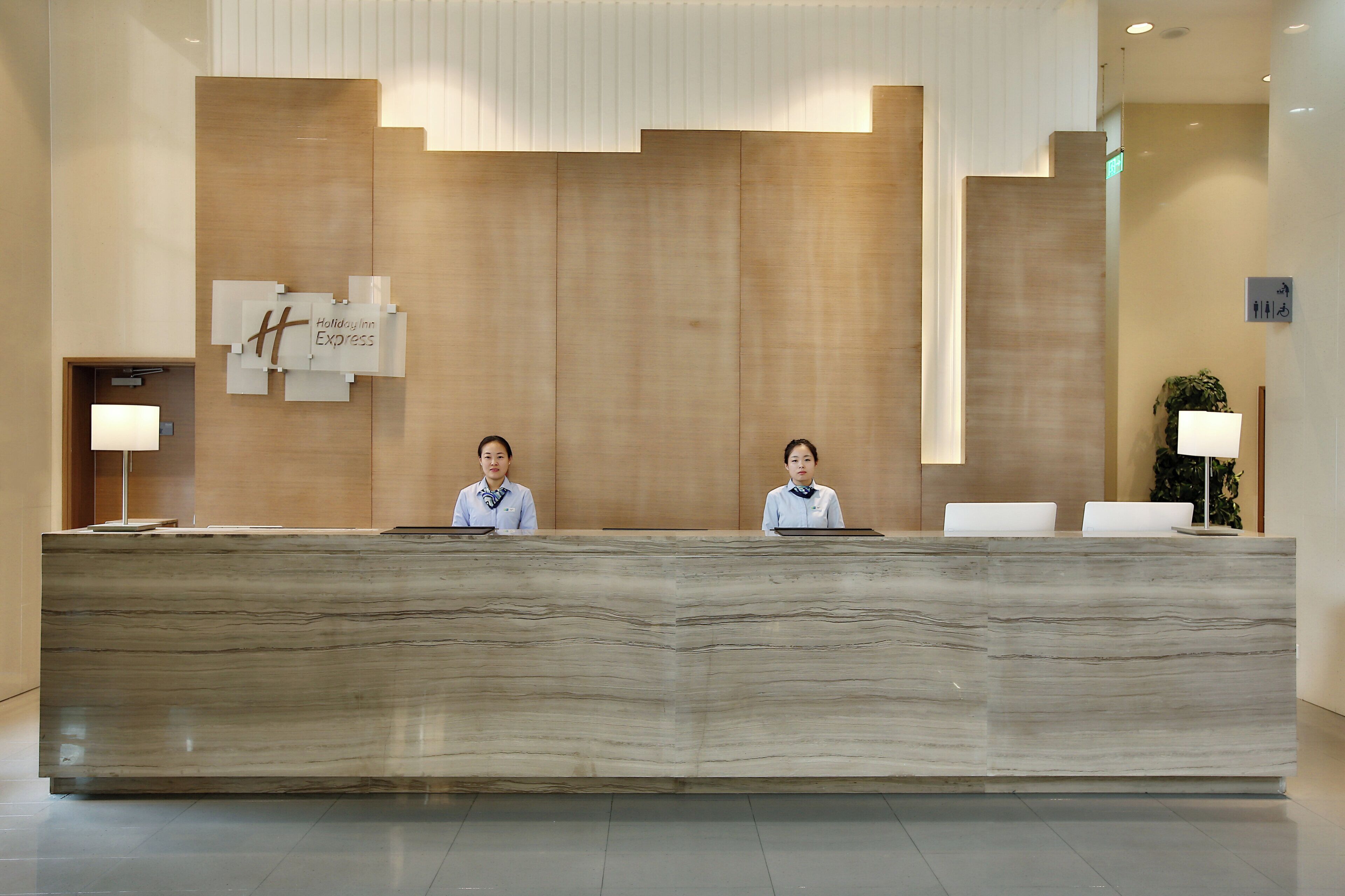 A reception desk with two staff members, a wooden wall with a logo, and a potted plant.