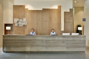 Un mostrador de recepción con dos empleados, una pared de madera con un logotipo y una planta en maceta.