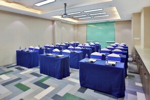 Un salón de conferencias con varias mesas, cada una con una botella de agua y una placa de identificación, y una pantalla de proyector al frente.