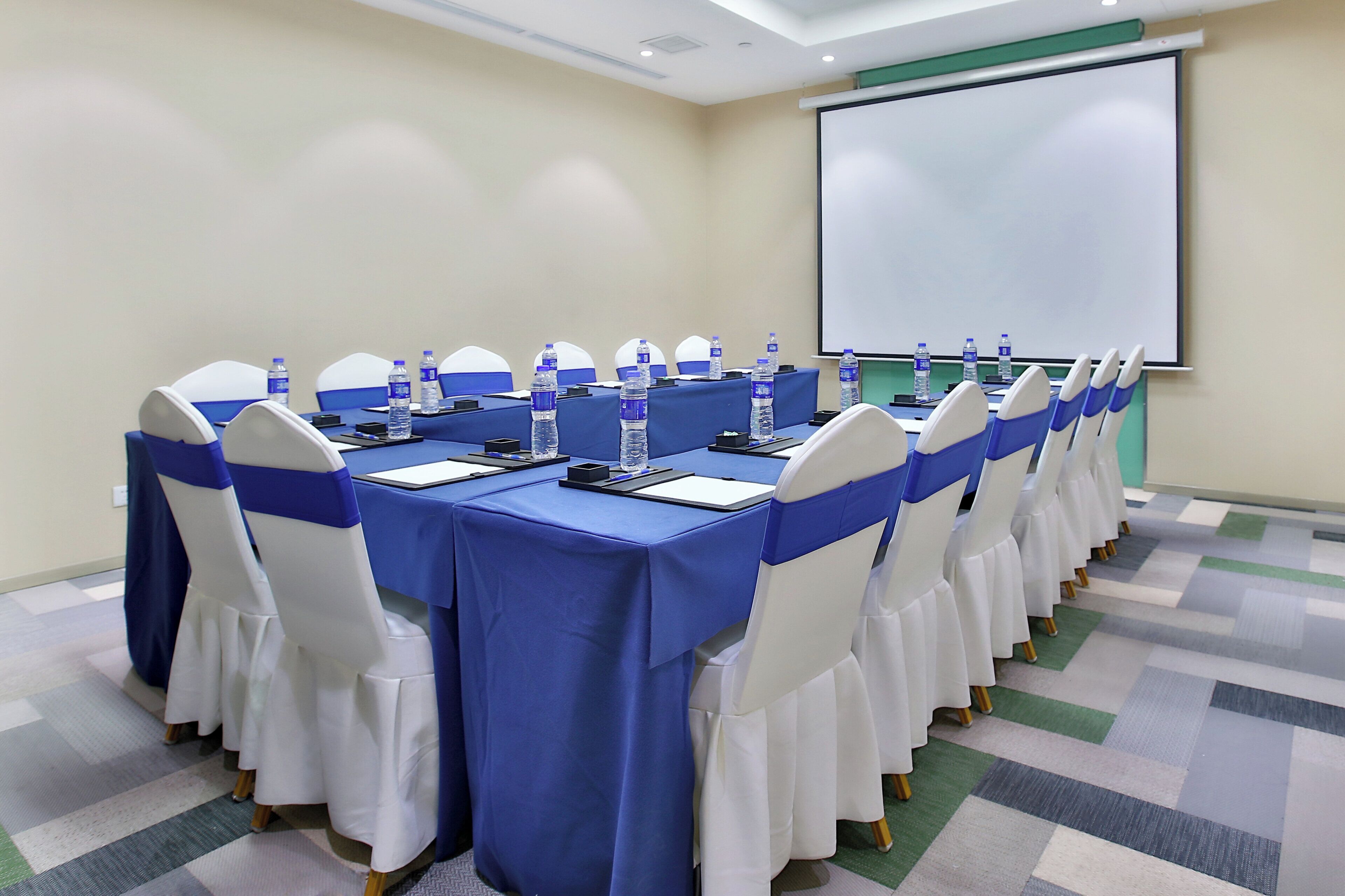 A conference room with a long table, chairs with blue sashes, water bottles, and a projector screen.