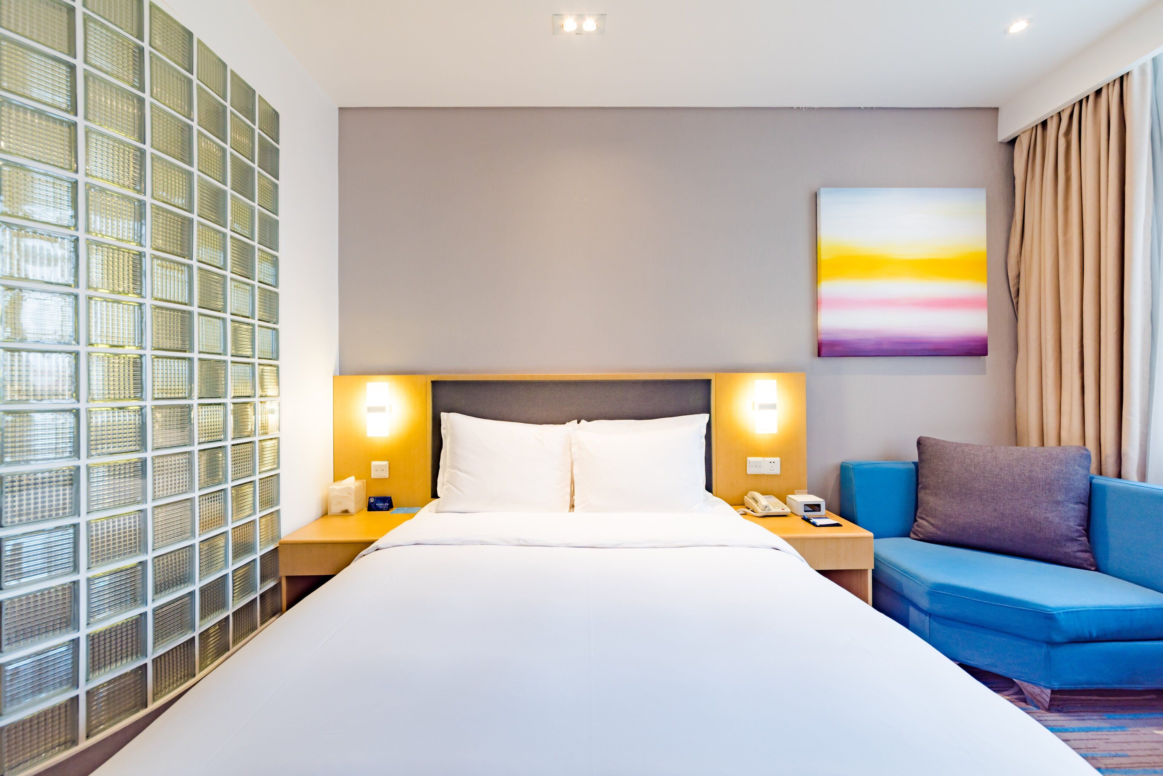 Une chambre d’hôtel avec un grand lit, deux tables de chevet avec des lampes, un canapé bleu et une cloison en verre.