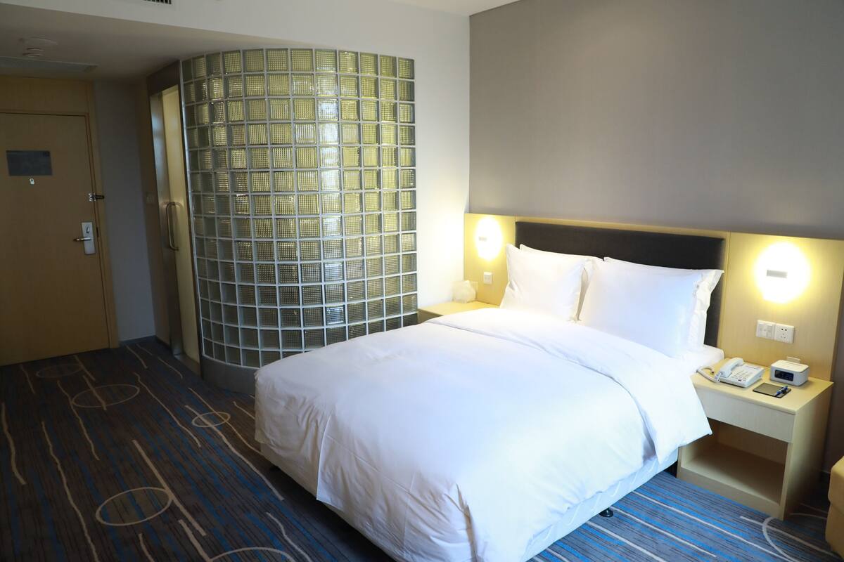 Une chambre d’hôtel avec un grand lit, une table de chevet avec un téléphone et une lampe, un luminaire fixé au mur et une porte avec un panneau en verre.