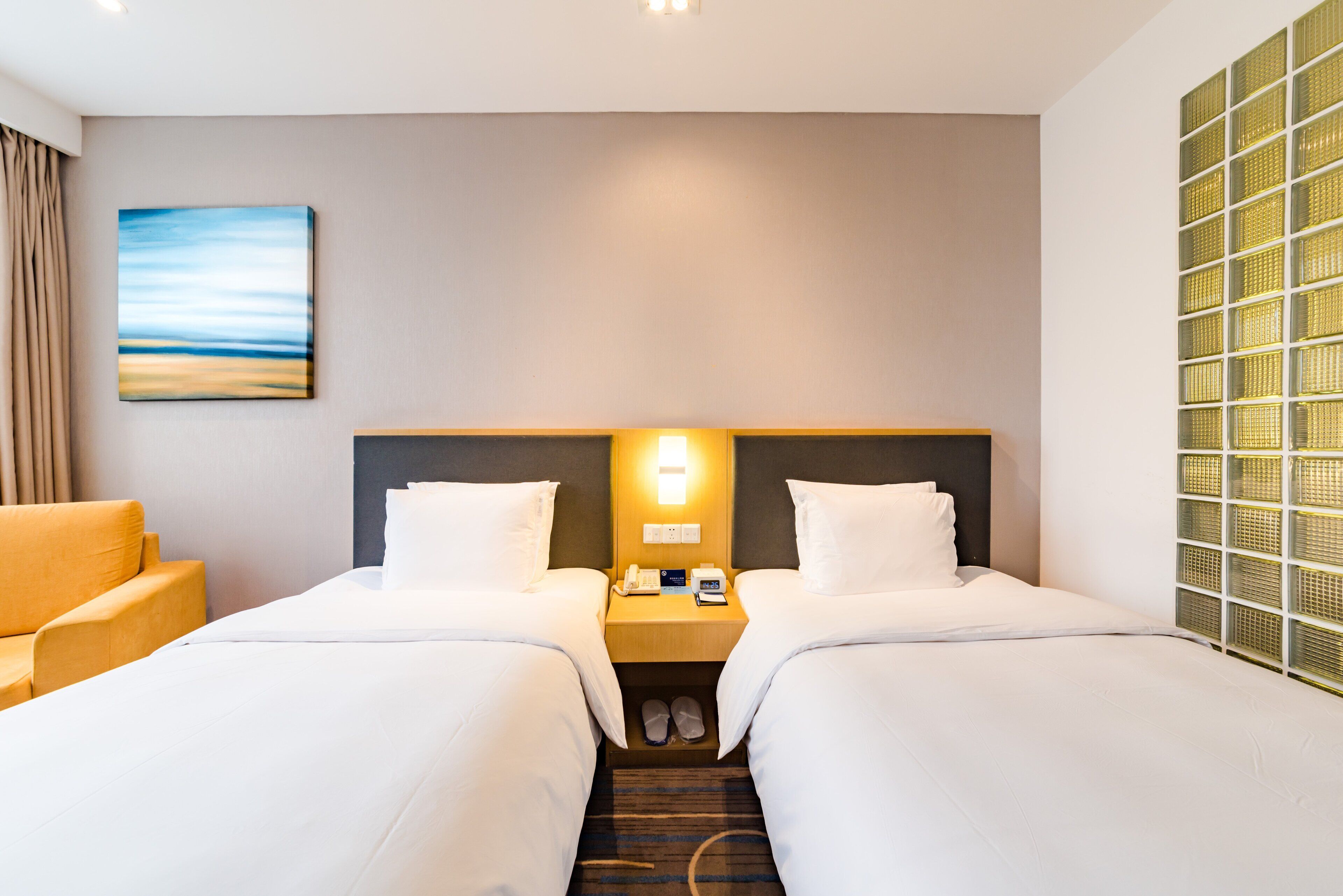 A hotel room with two beds, a nightstand with a phone and lamp, a pair of slippers, and a painting on the wall.