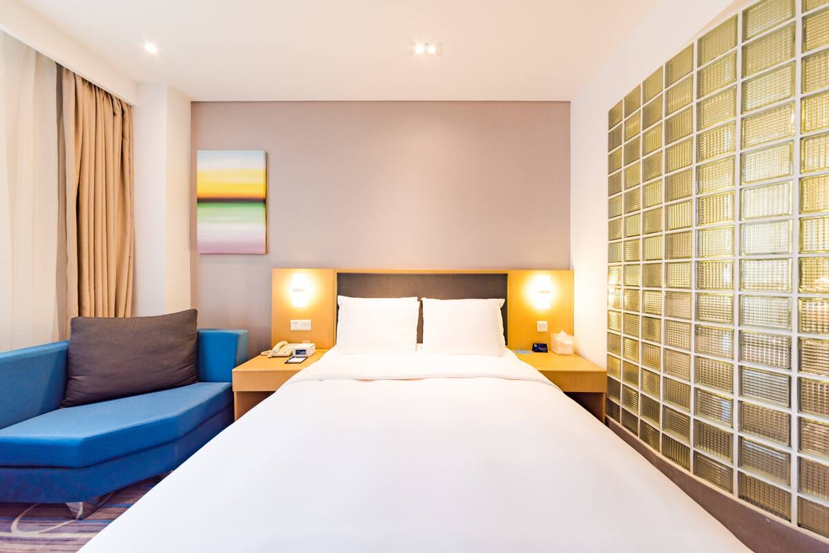 Une chambre d’hôtel avec un grand lit, un canapé bleu, deux tables de chevet avec des lampes, un tableau au mur et un mur décoratif en verre.