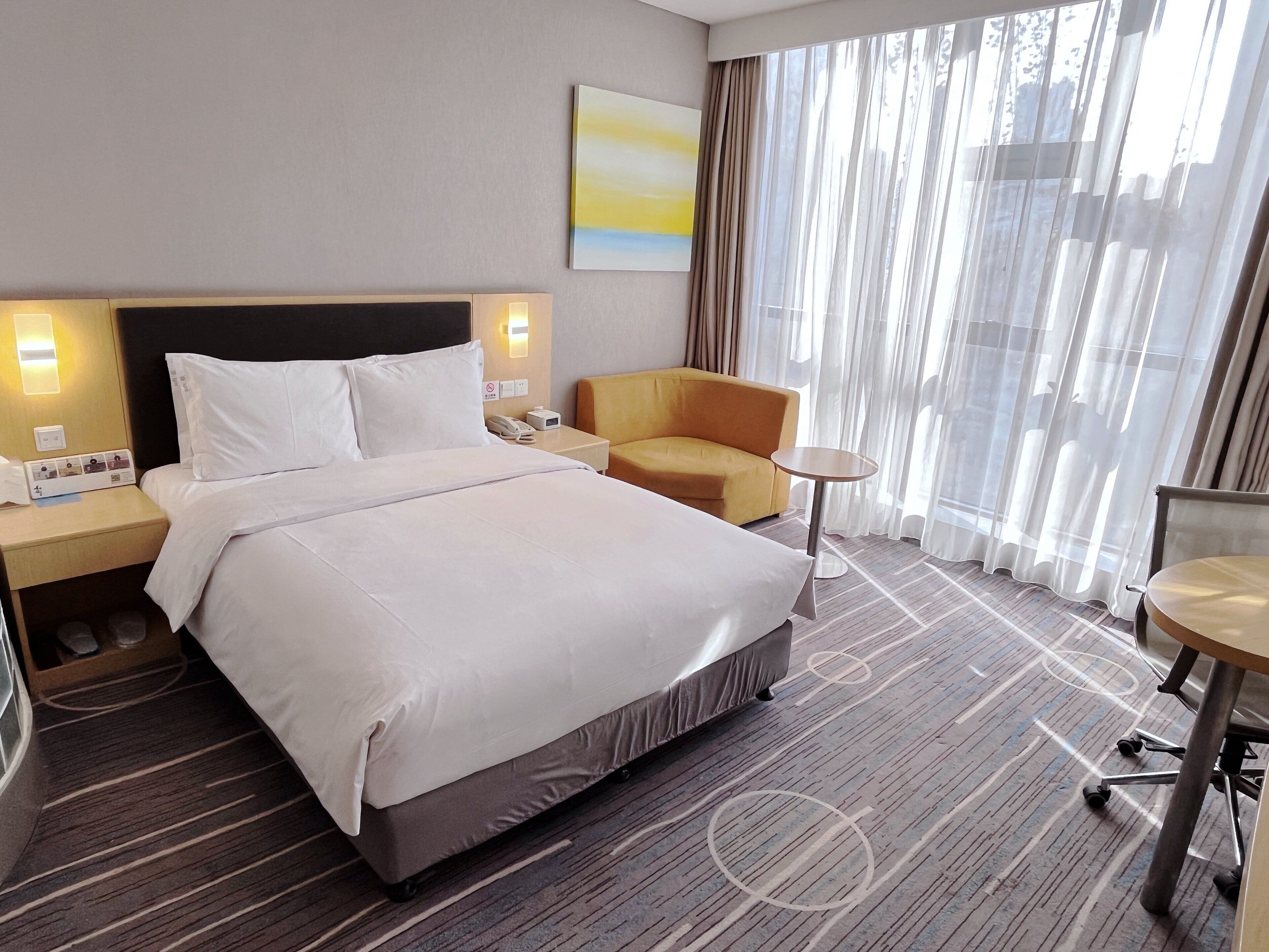Ein Hotelzimmer mit einem großen Bett, einem Nachttisch mit Telefon, einem kleinen runden Tisch, einem gelben Sessel und Blick ins Freie durch durchsichtigen Vorhängen.