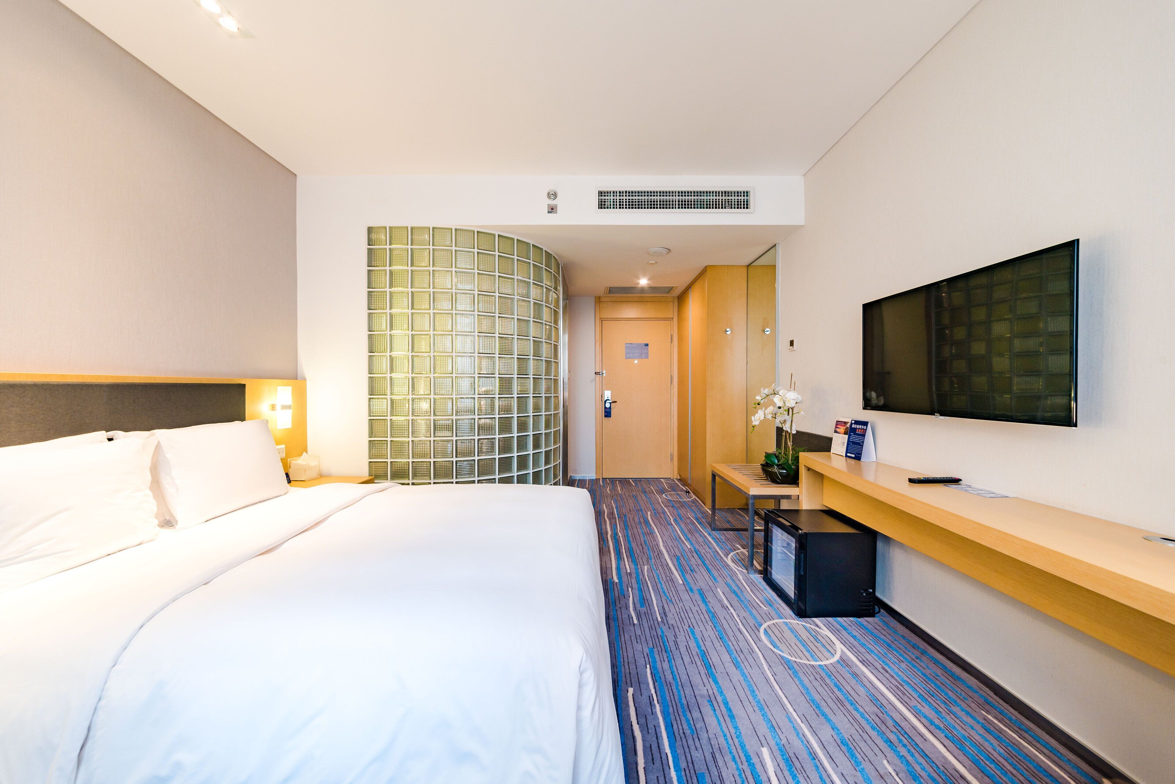 Ein Hotelzimmer mit einem großen Bett, einem Schreibtisch, einem Flachbildfernseher, einem Spiegel und einem gemusterten Teppich.