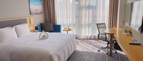 A hotel room with a large bed, a desk, a chair, a small table, and a large window with curtains.