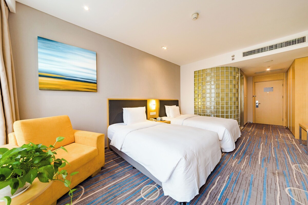 Ein Hotelzimmer mit zwei Betten, einem Schlafsofa, einem Gemälde und einem großen Fenster.