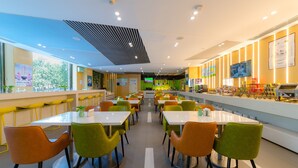 A modern cafeteria with multiple tables and chairs, a counter with food items, and a green screen in the background. - Holiday Inn Express Shijiazhuang Heping by IHG (Shijiazhuang)