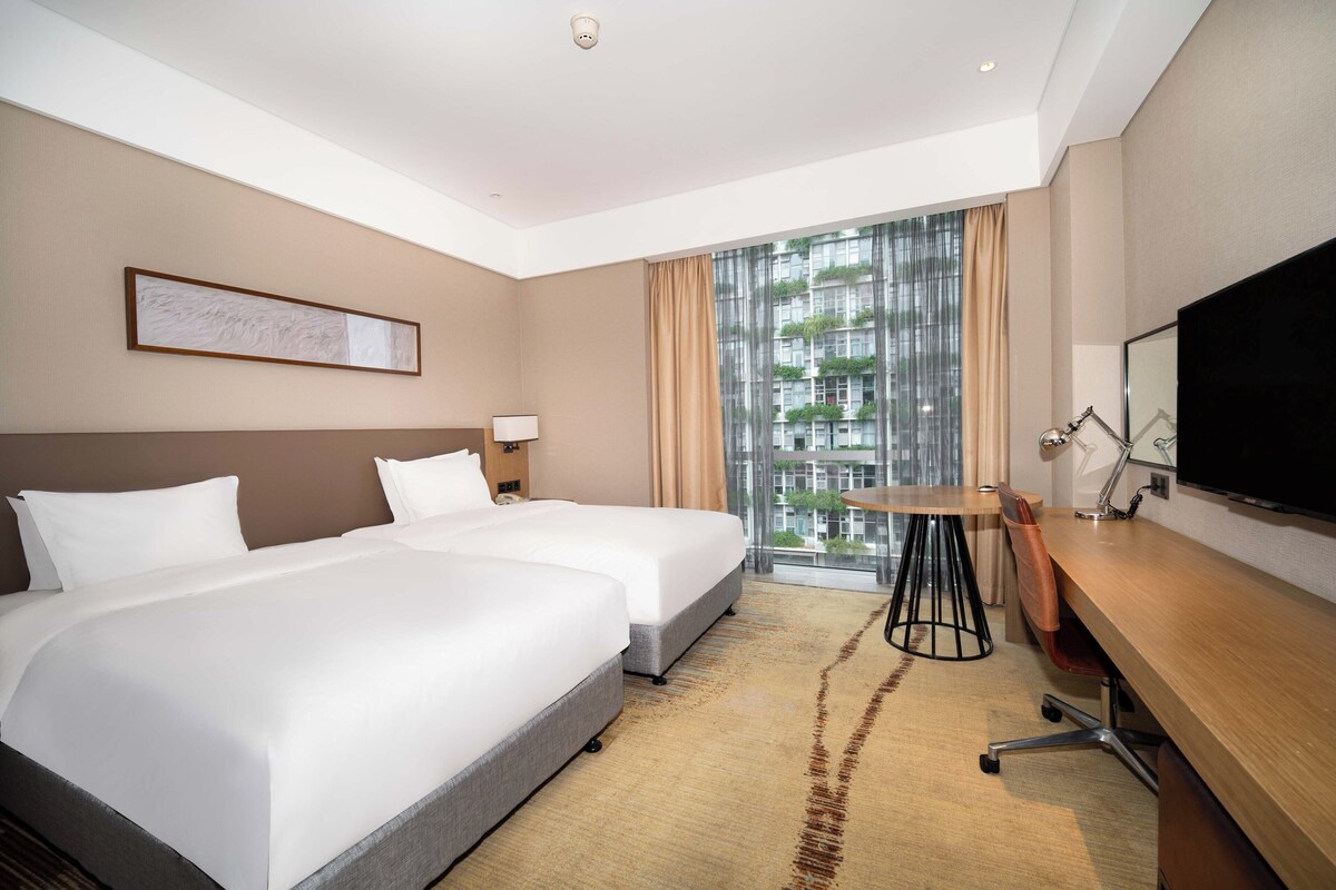 Een hotelkamer met twee bedden, een bureau, een televisie en een raam met uitzicht op gebouwen.