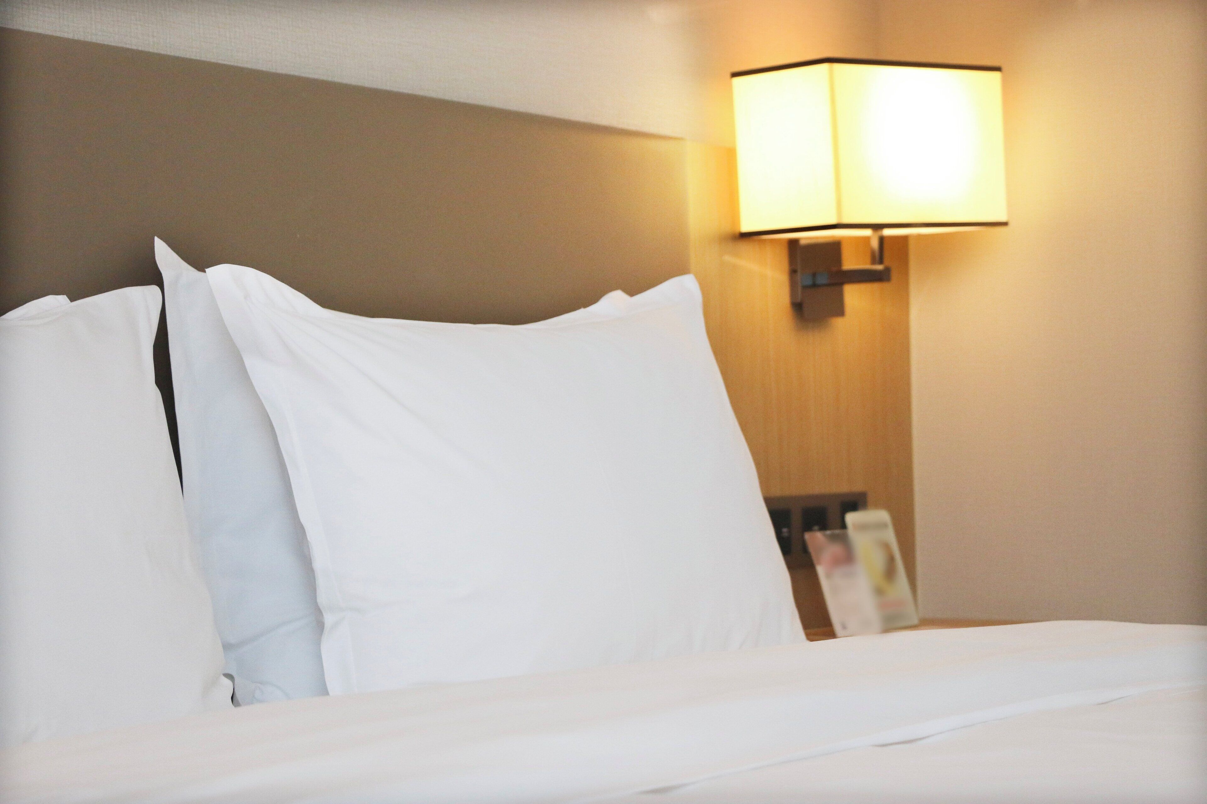 Una habitación de hotel con una cama bien hecha, una lámpara de pared y una mesita de noche con una revista.