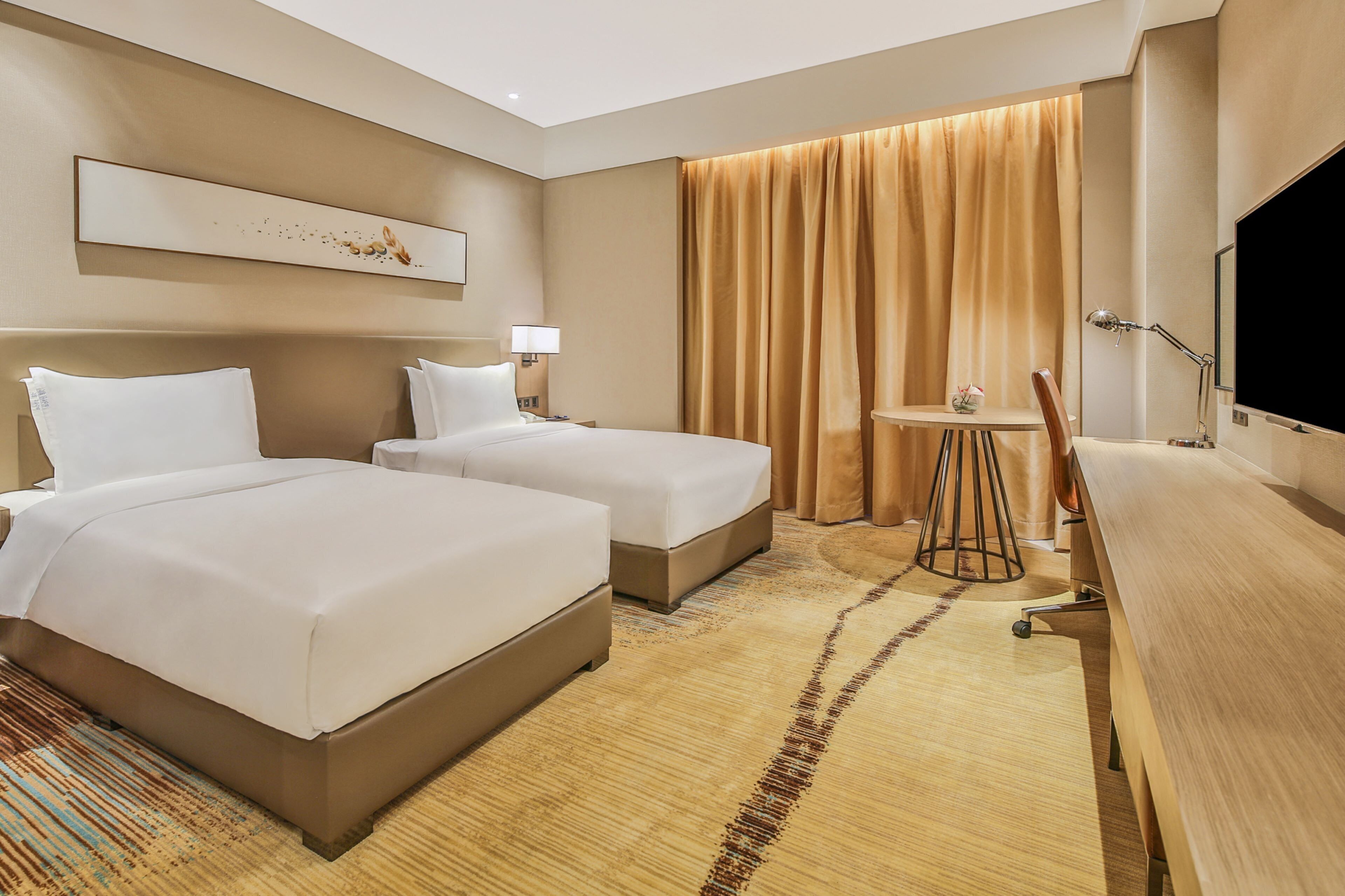 A hotel room with two beds, a desk, a chair, a TV, and a window with curtains.