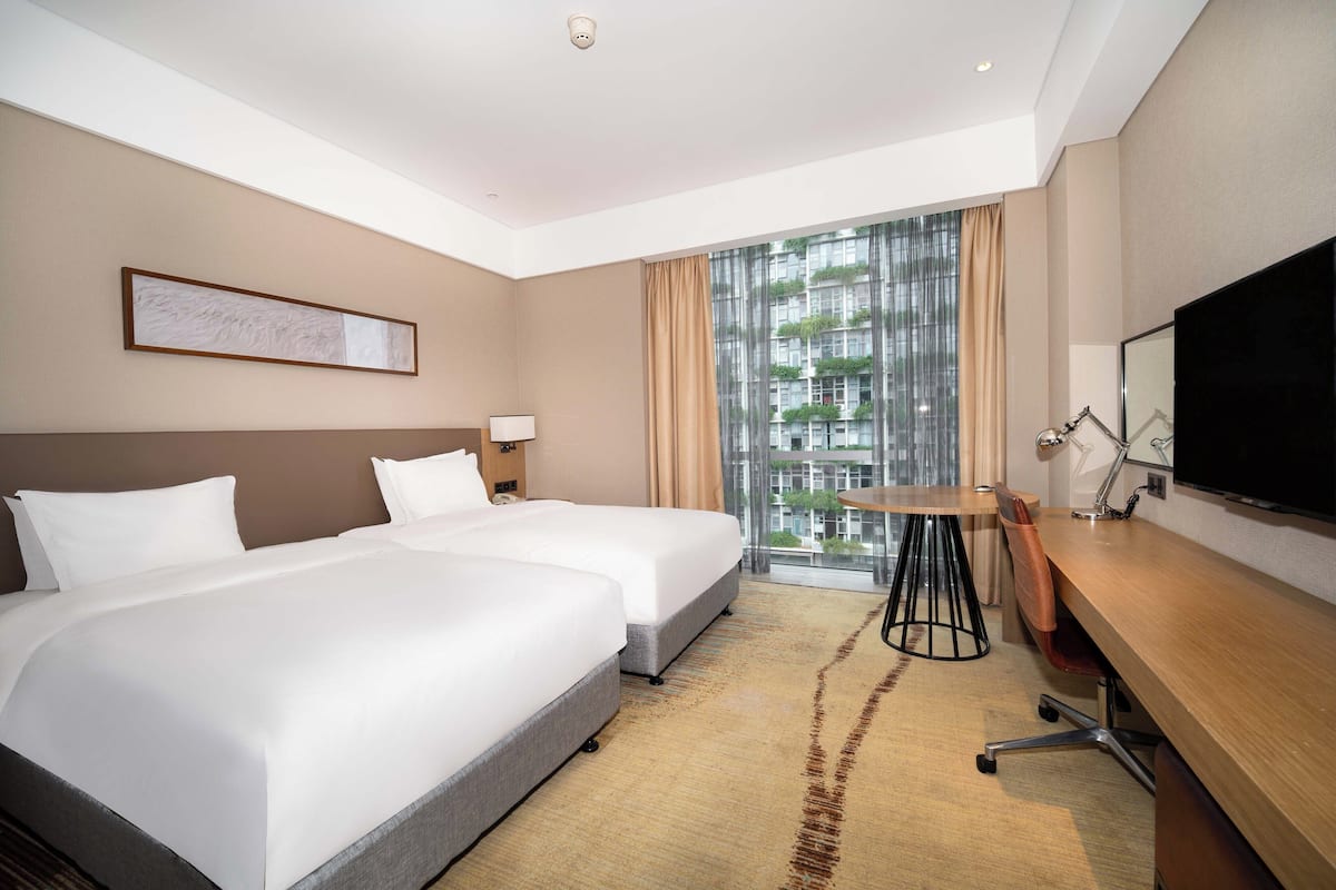 A hotel room with two beds, a desk, a TV, and a window with a view of buildings.