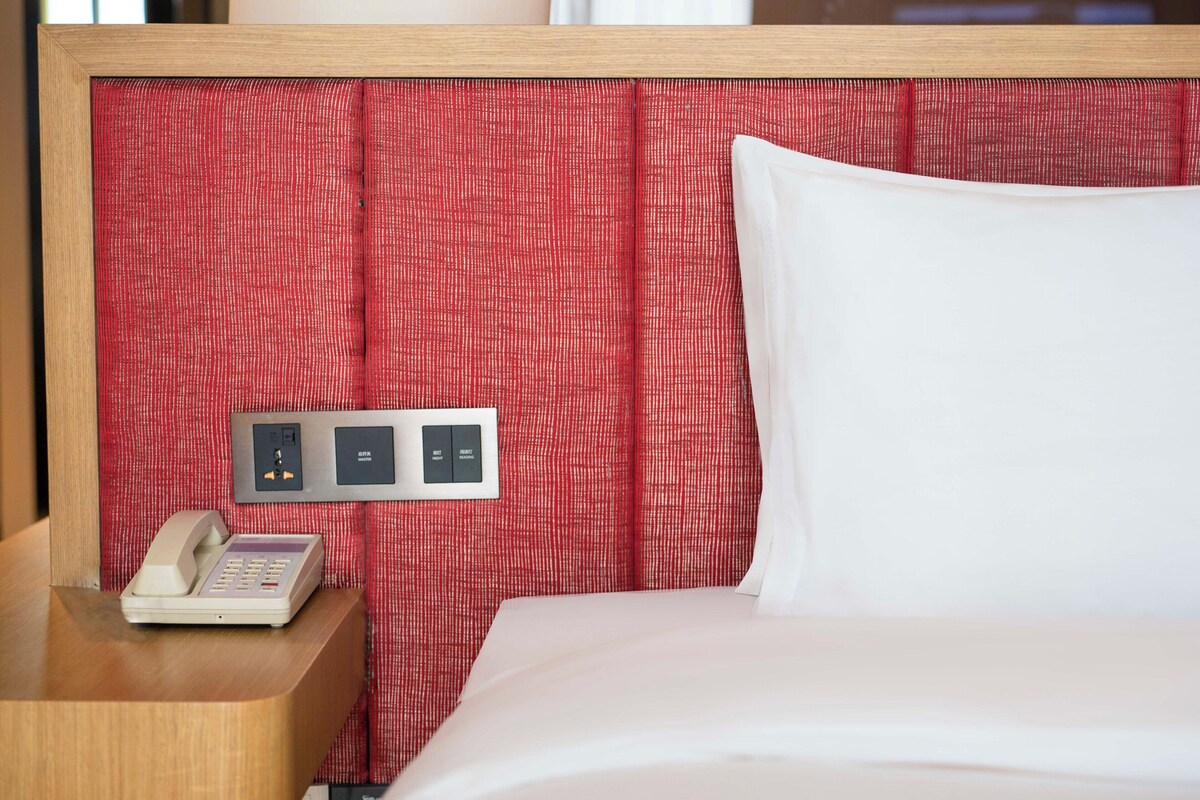 Hotelkamer met een houten hoofdbord, een rood gordijn met patroon, een wit kussen en een telefoon op het nachtkastje.
