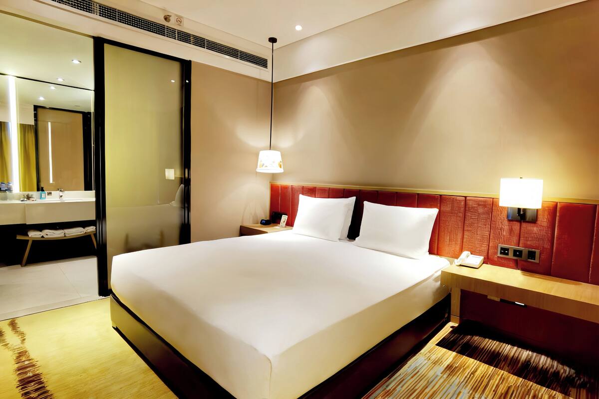 A hotel room with a large bed, a bedside table with a lamp, a nightstand, a mirror, and a bathroom with a sink and towel rack.
