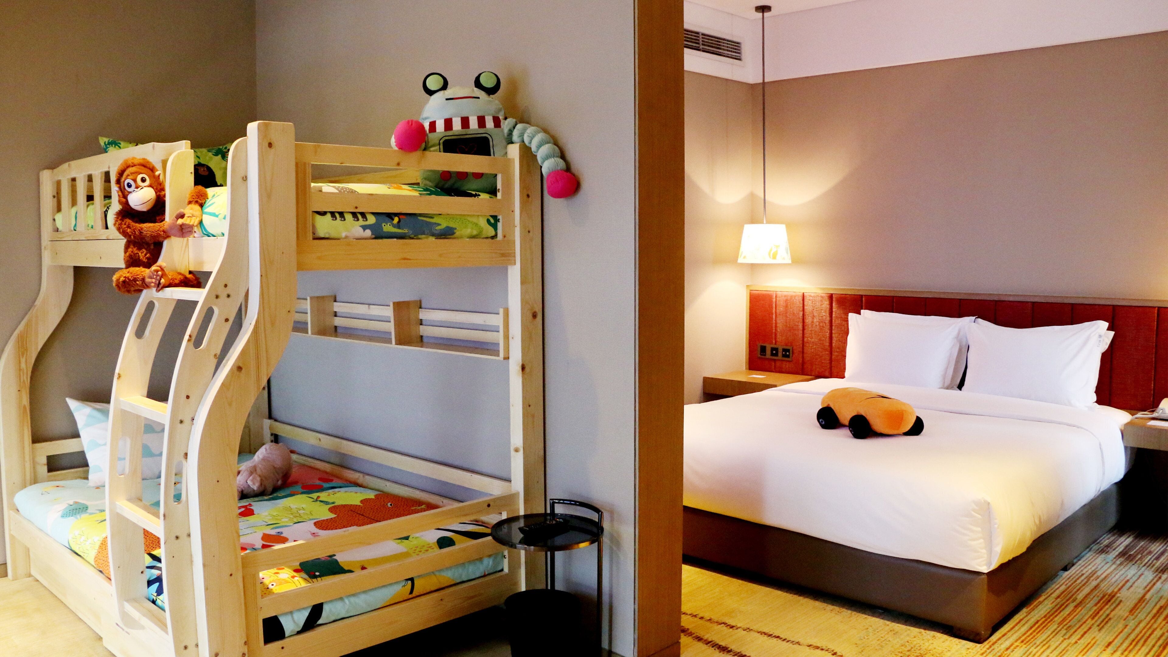 Una cama litera con un monito de juguete y un peluche en la parte superior, una cama individual con un almohadón y un coche de juguete, y una mesita de noche con una lámpara.