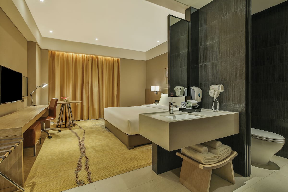 A modern hotel room with a large bed, a desk, a desk lamp, a chair, a bathroom with a sink and toilet, and a TV.