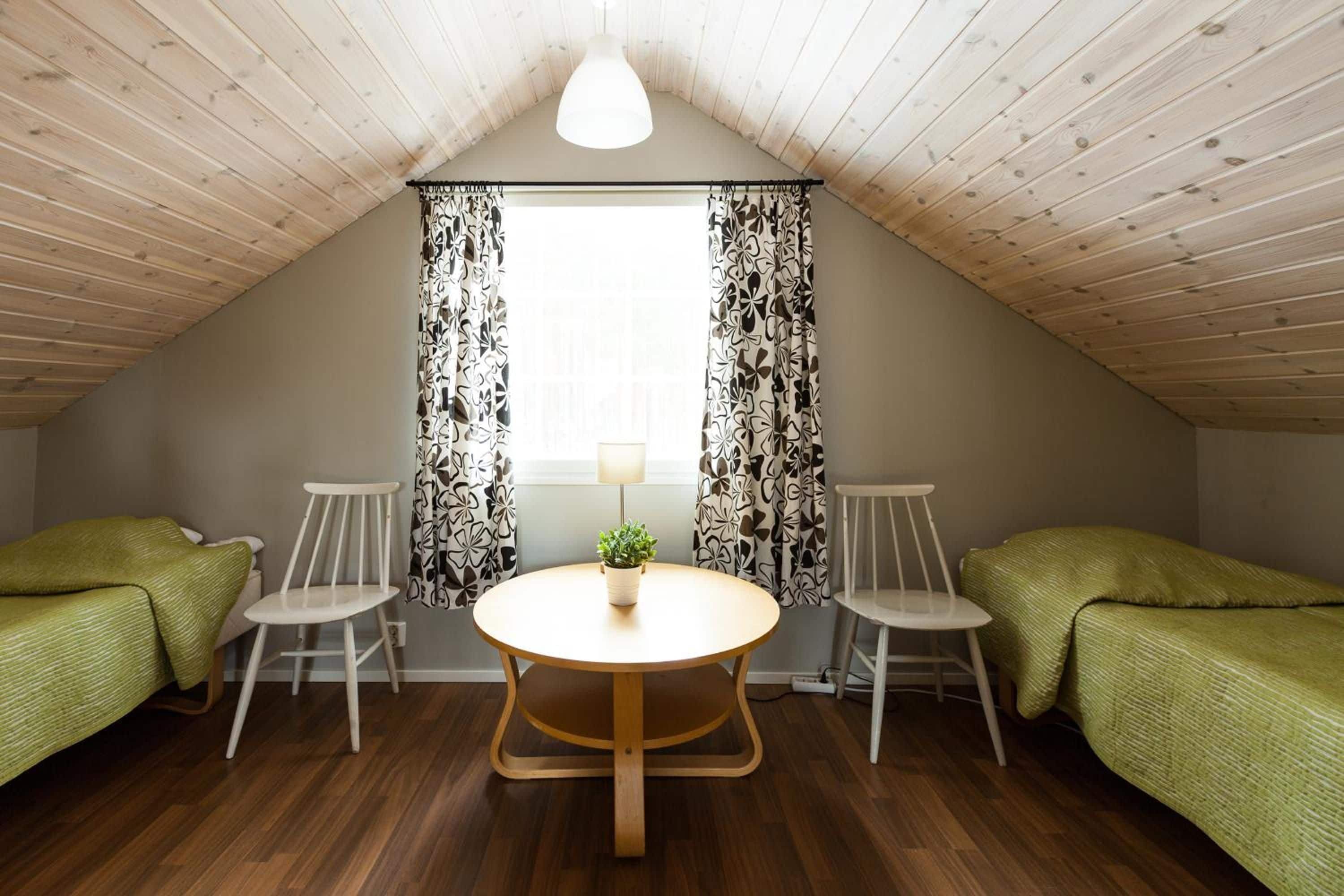 A small, cozy room with a sloped ceiling, two single beds, a round table with chairs, and a window with patterned curtains.