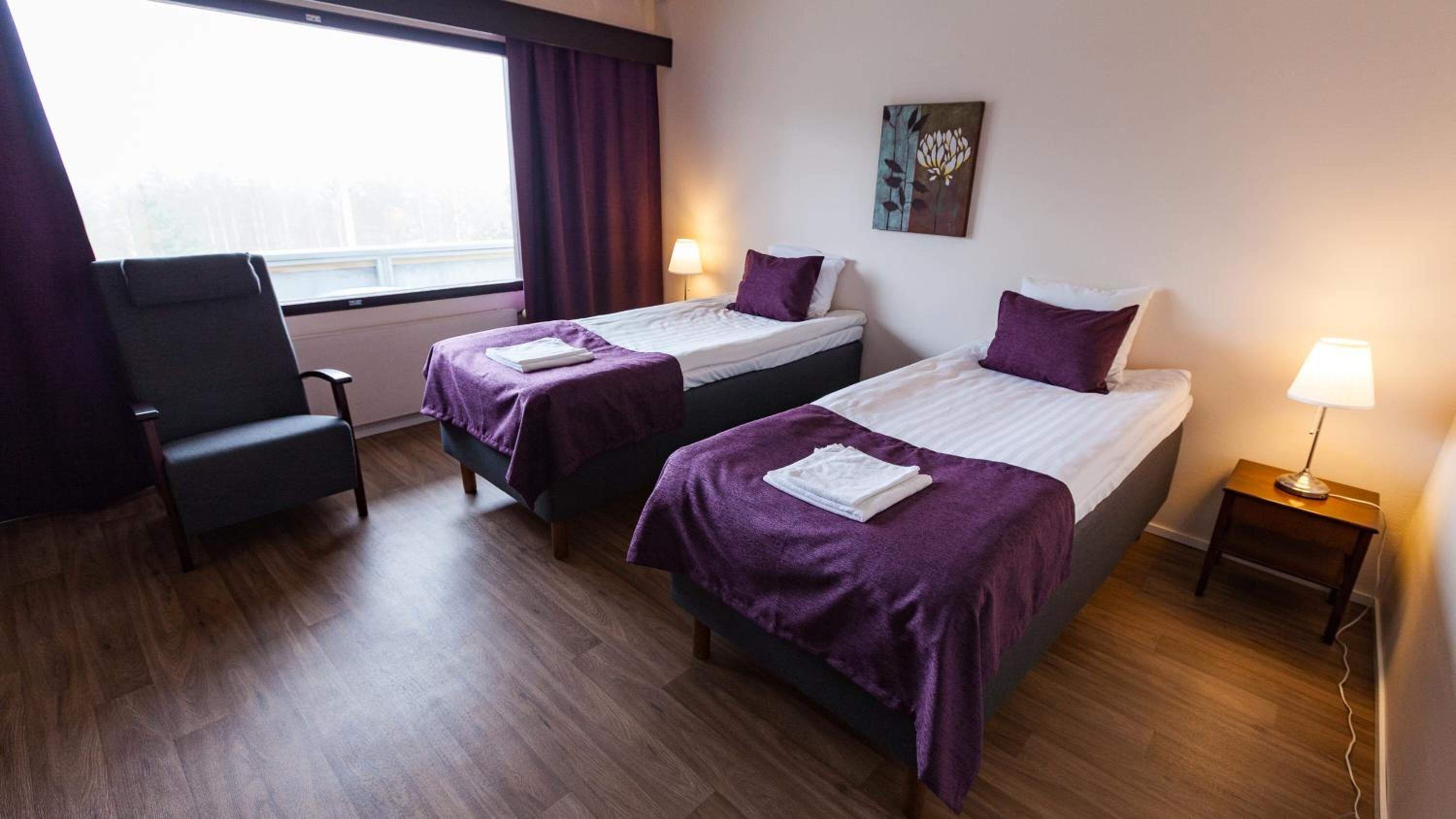 A hotel room with two single beds, a chair, a bedside table with a lamp, and a window with curtains.