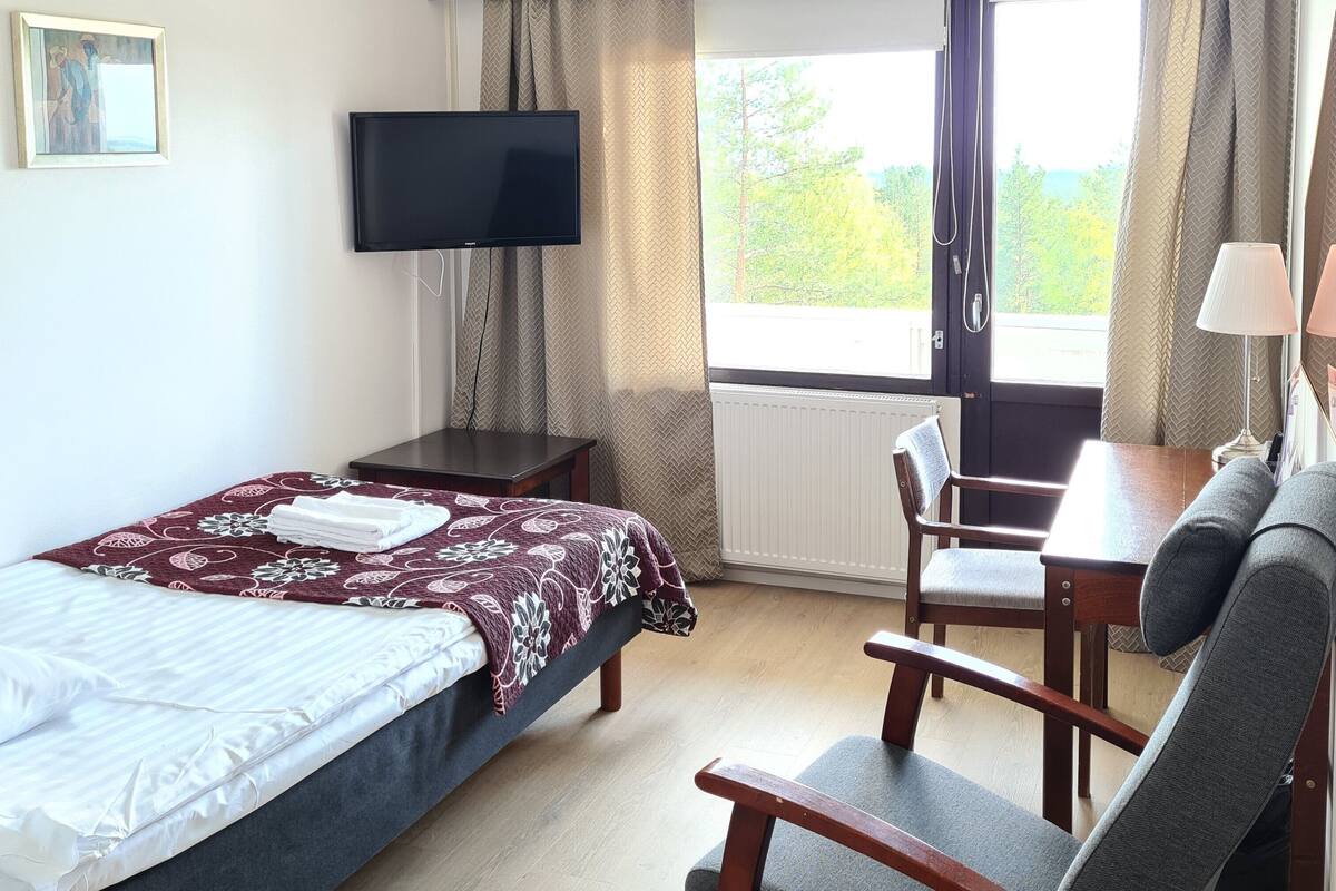 A hotel room with a bed, a desk, a chair, a TV, and a window with curtains.