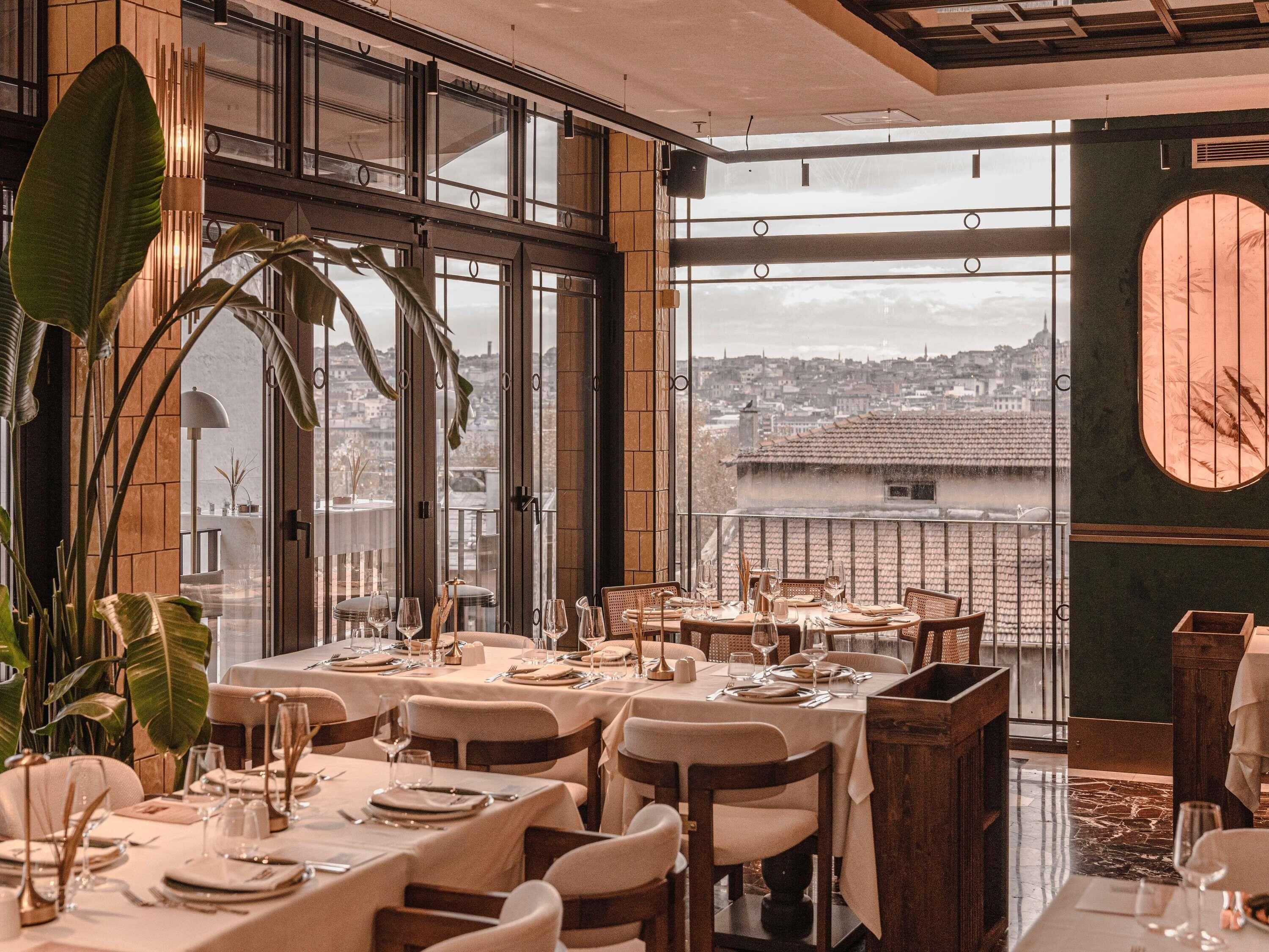 Ein Restaurant mit gedeckten Tischen, großen Fenstern mit Blick auf die Stadt und Zimmerpflanzen.