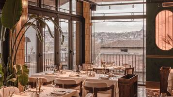 Ein Restaurant mit gedeckten Tischen, großen Fenstern mit Blick auf die Stadt und Zimmerpflanzen.