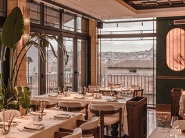 Un restaurante con mesas dispuestas para comer, grandes ventanales con vistas a la ciudad y plantas de interior.
