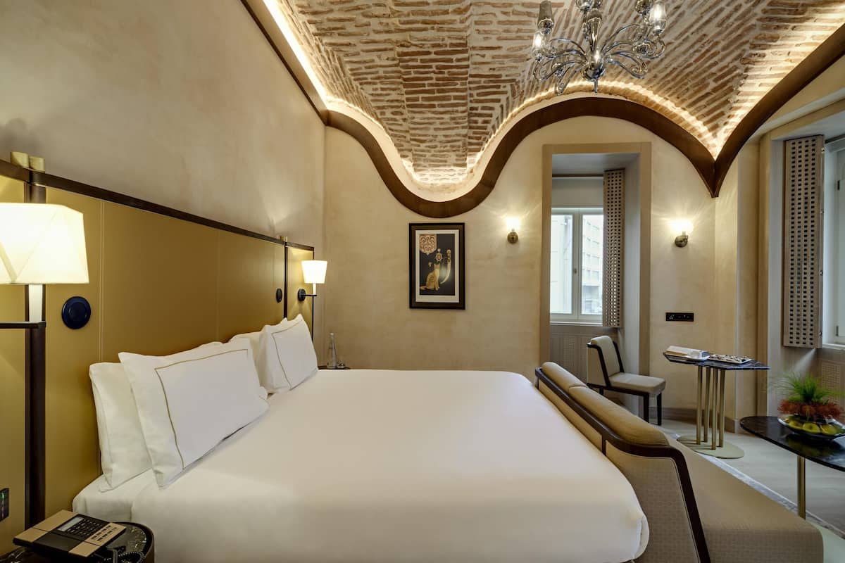 Ein Hotelzimmer mit einem großen Bett, einem Schreibtisch mit Telefon, einem Stuhl, einem kleinen Tisch mit einer Vase und einem Bild an der Wand.