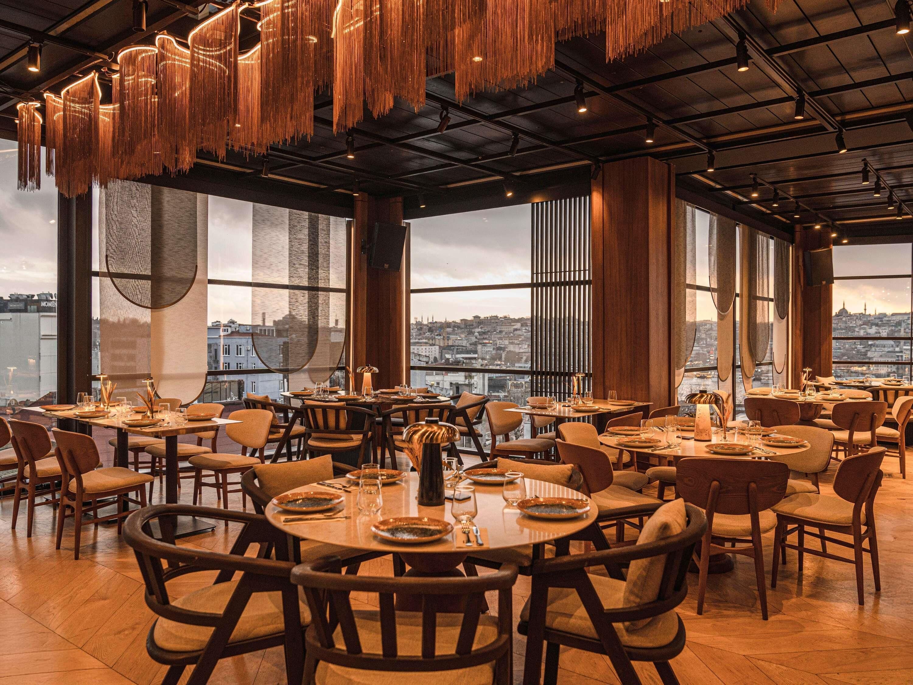 Un restaurant avec des tables et des chaises rondes, ainsi que des luminaires suspendus. De grandes fenêtres offrent une vue sur la ville.