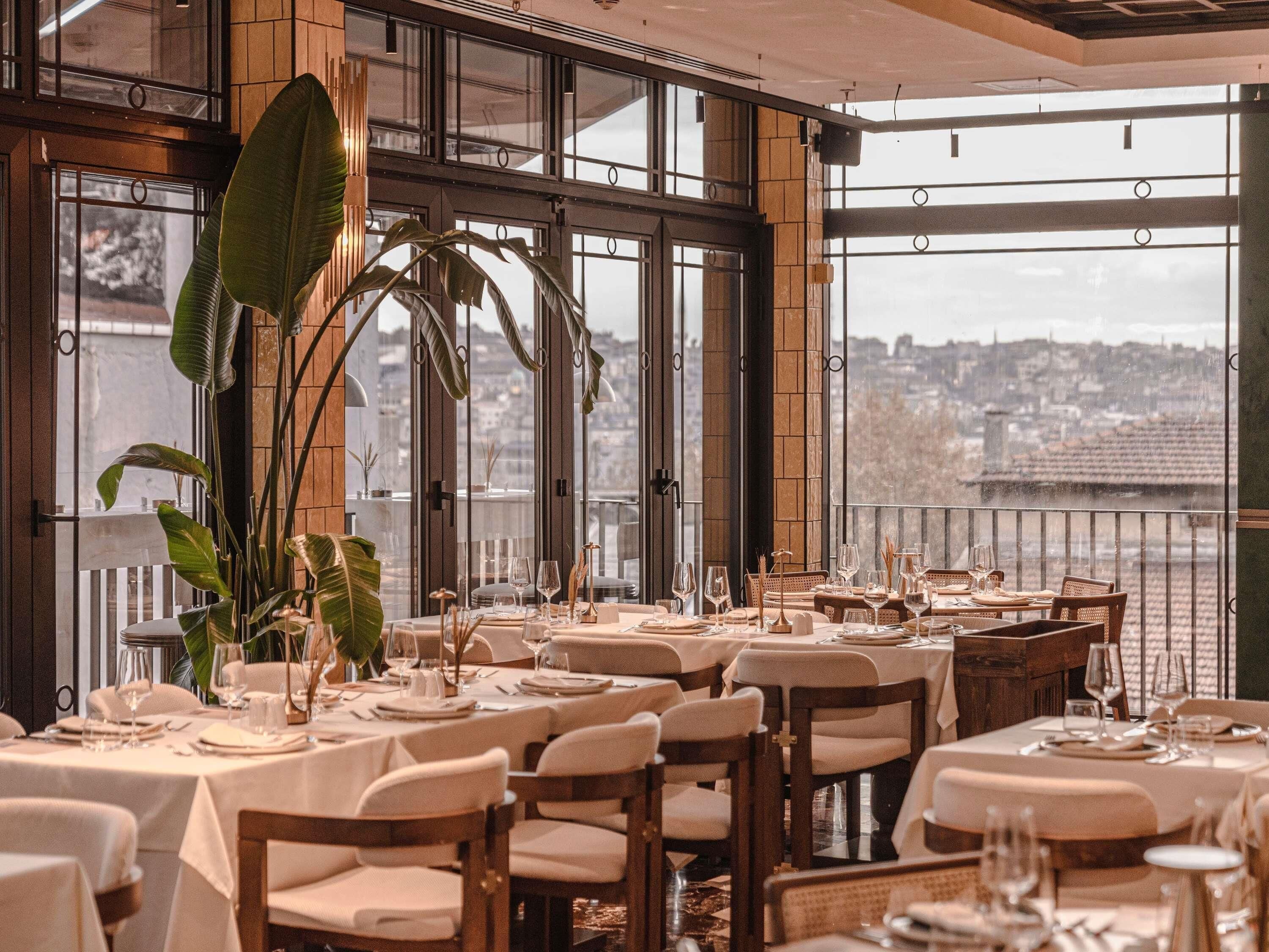 Un ristorante con diversi tavoli apparecchiati, ampie finestre che si affacciano sulla città e piante in vaso.