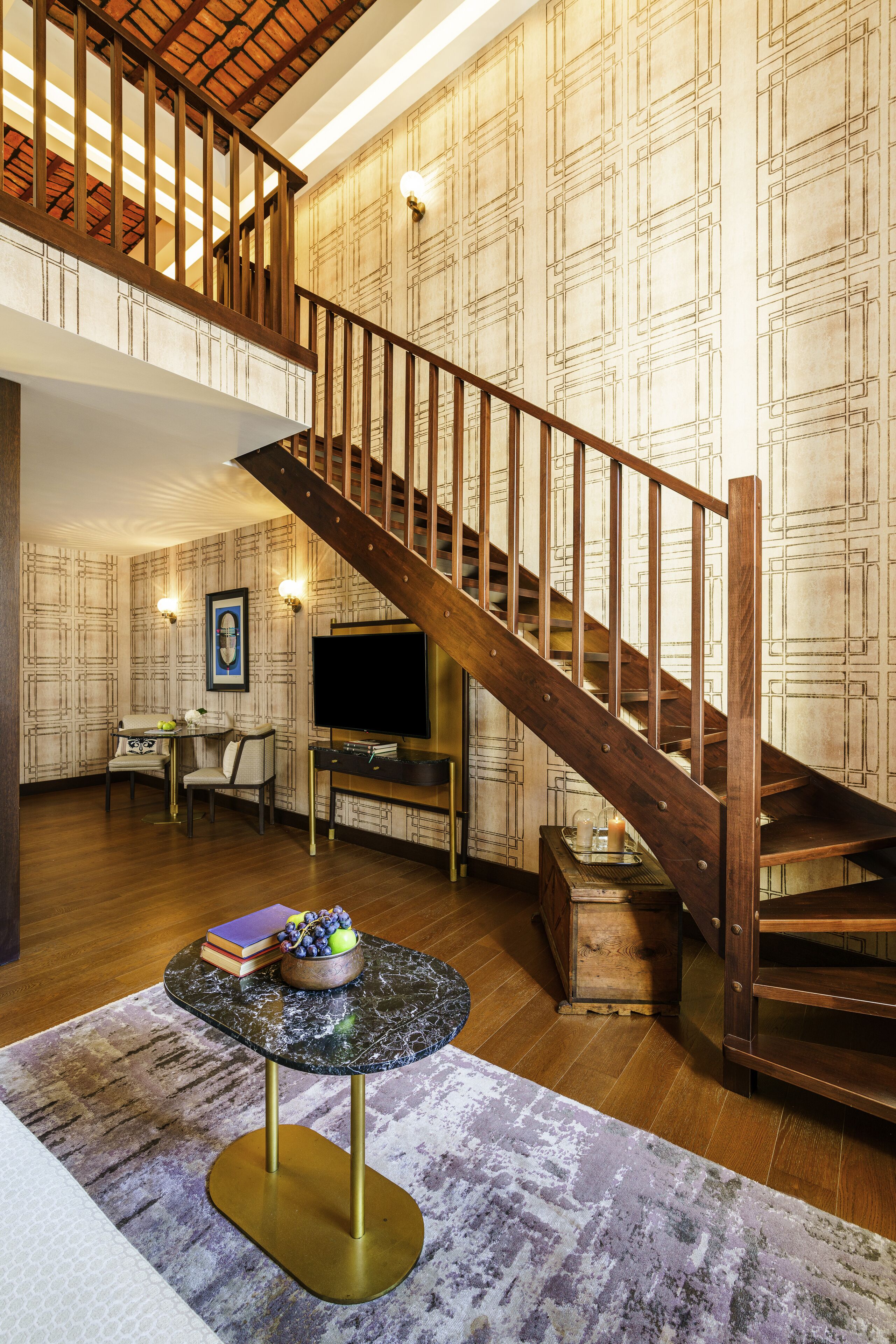 A spacious living area with a wooden staircase, a flat-screen TV, a dining table with chairs, and a decorative bowl on a marble table.