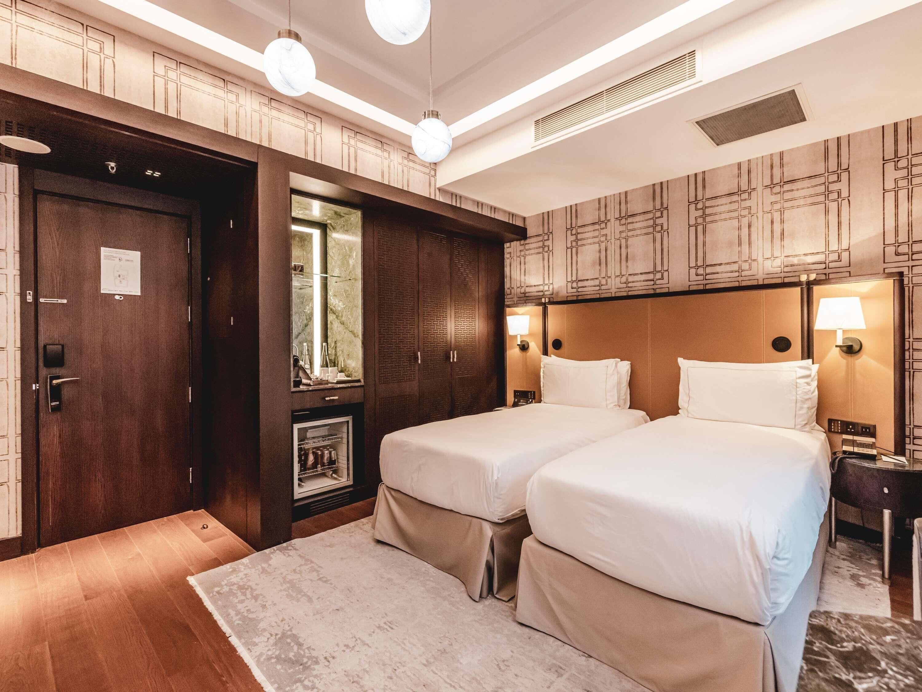 A hotel room with two beds, a wooden wardrobe, a mirror, and a nightstand with a lamp.