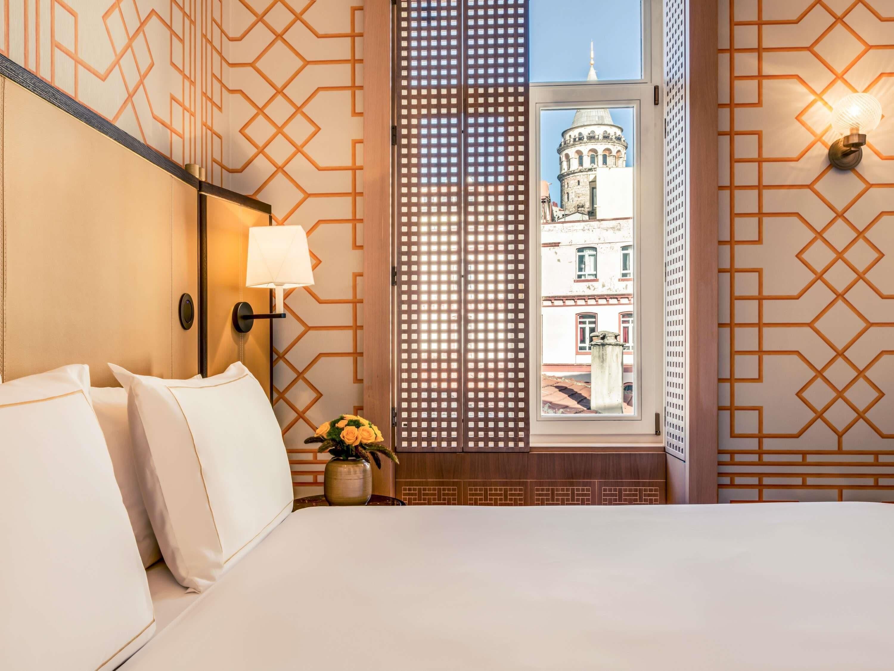 A hotel room with a bed, a nightstand with a lamp, a vase with flowers, a window with a view of a building, and patterned walls.