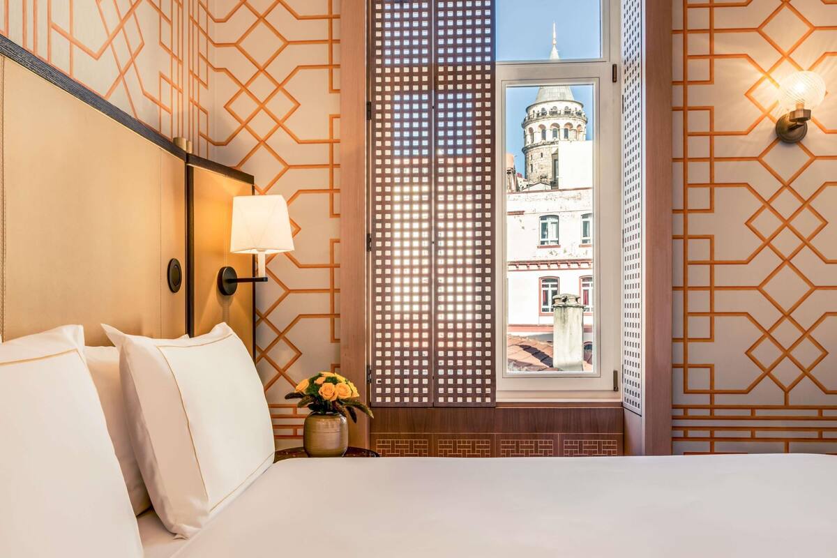 Ein Hotelzimmer mit einem Bett, einem Nachttisch mit Lampe, einer Vase mit Blumen, einem Fenster mit Blick auf ein Gebäude und Wänden mit Muster.
