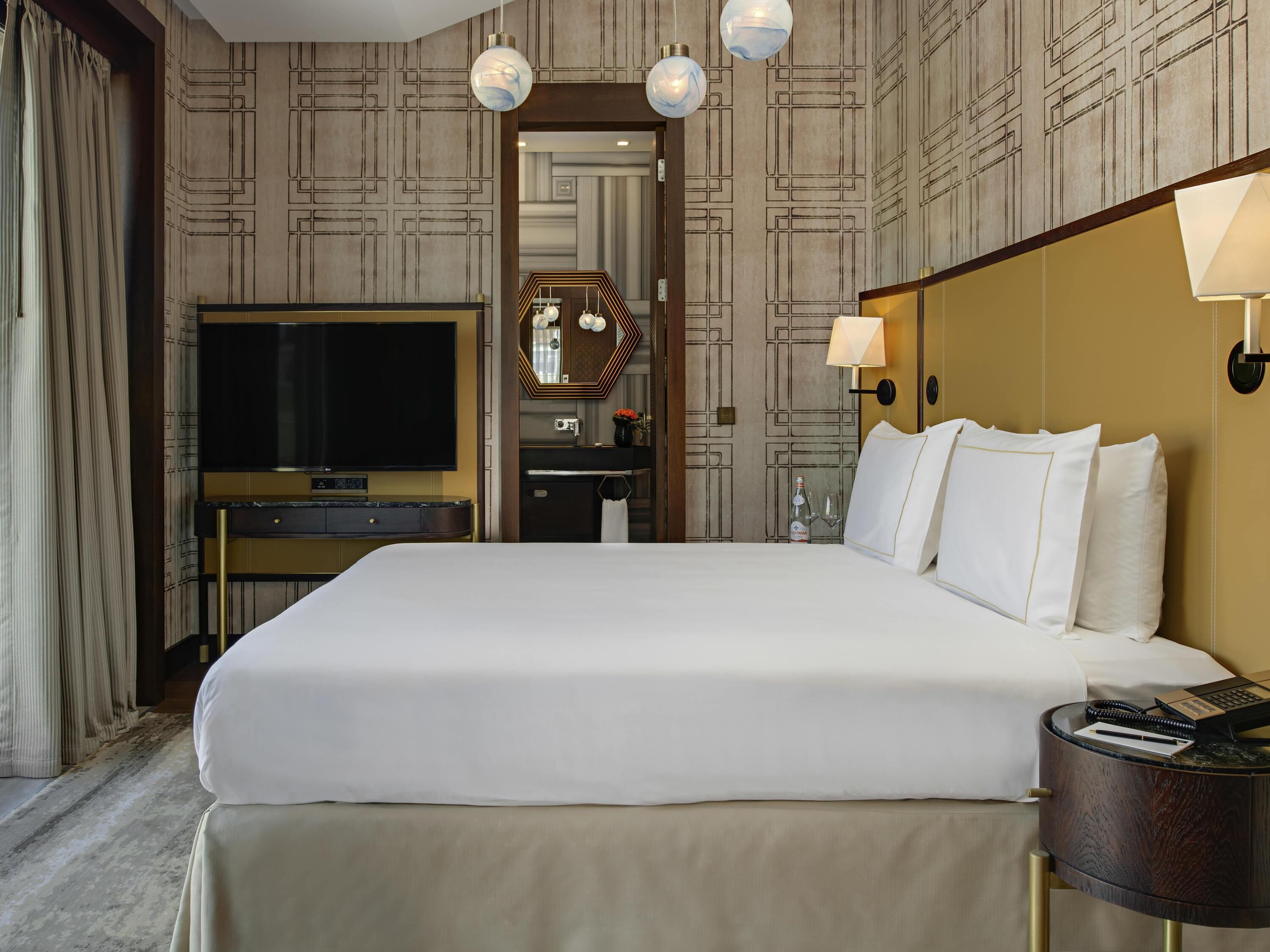 Habitación de hotel con una cama grande, televisión, minibar y baño visible a través de una puerta abierta.