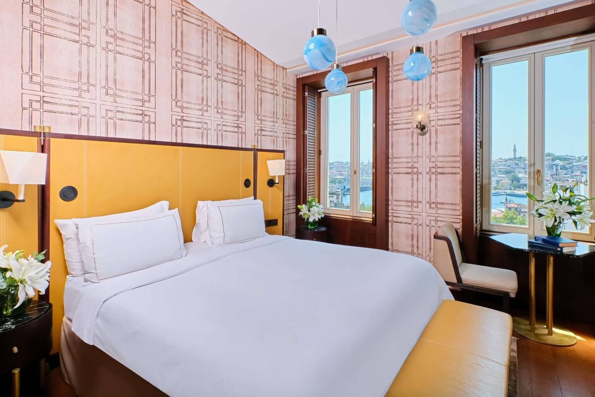 Ein Hotelzimmer mit einem großen Bett, einem Sessel, einem kleinen Tisch und Blick auf eine Stadtlandschaft durch große Fenster.