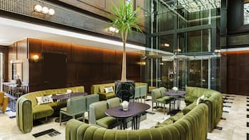 Una hall moderna con sedute rivestite in verde, una palma al centro e un ascensore in vetro.