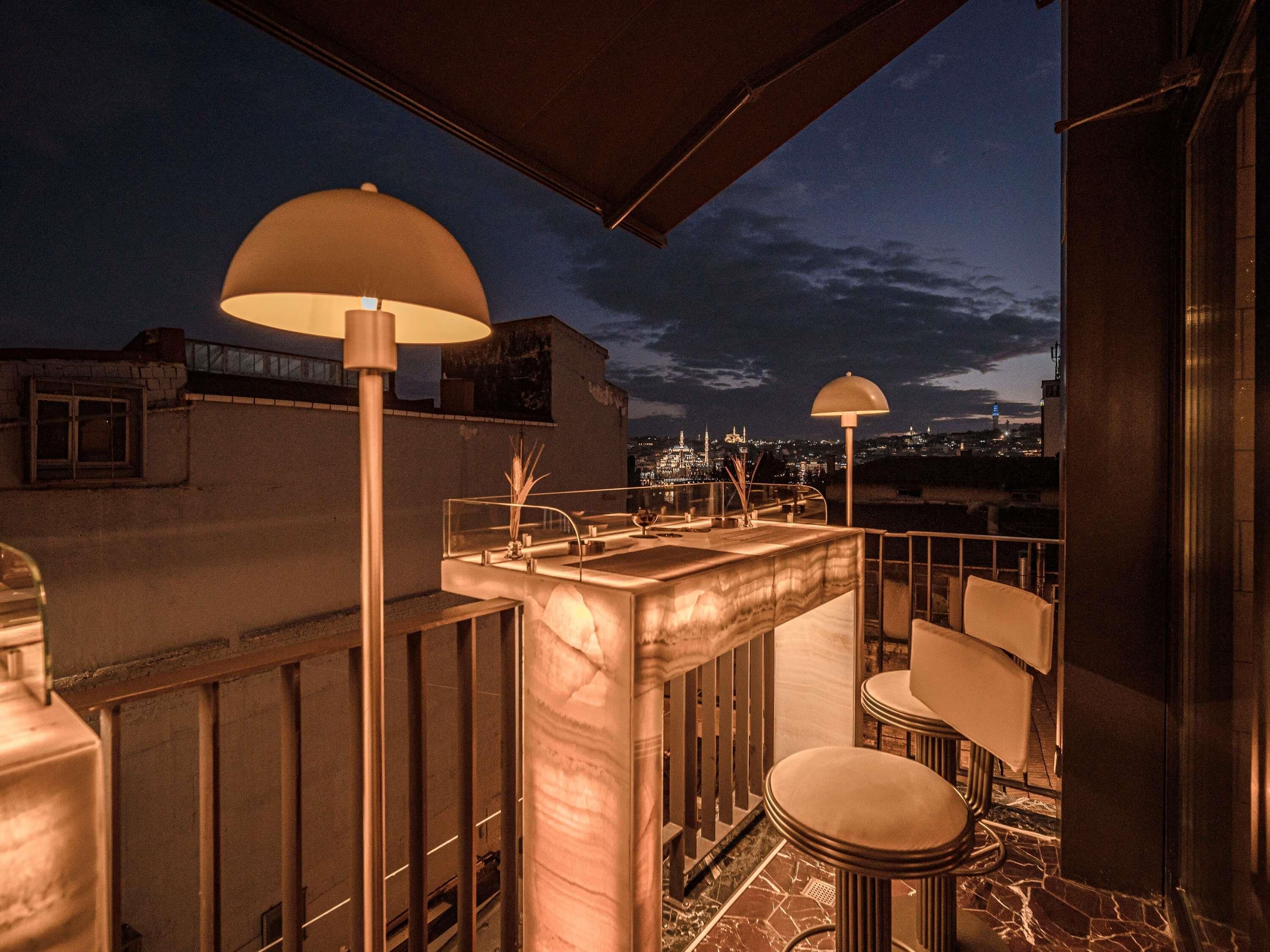 A rooftop terrace with a bar area, two large lamps, and a cityscape view at dusk.
