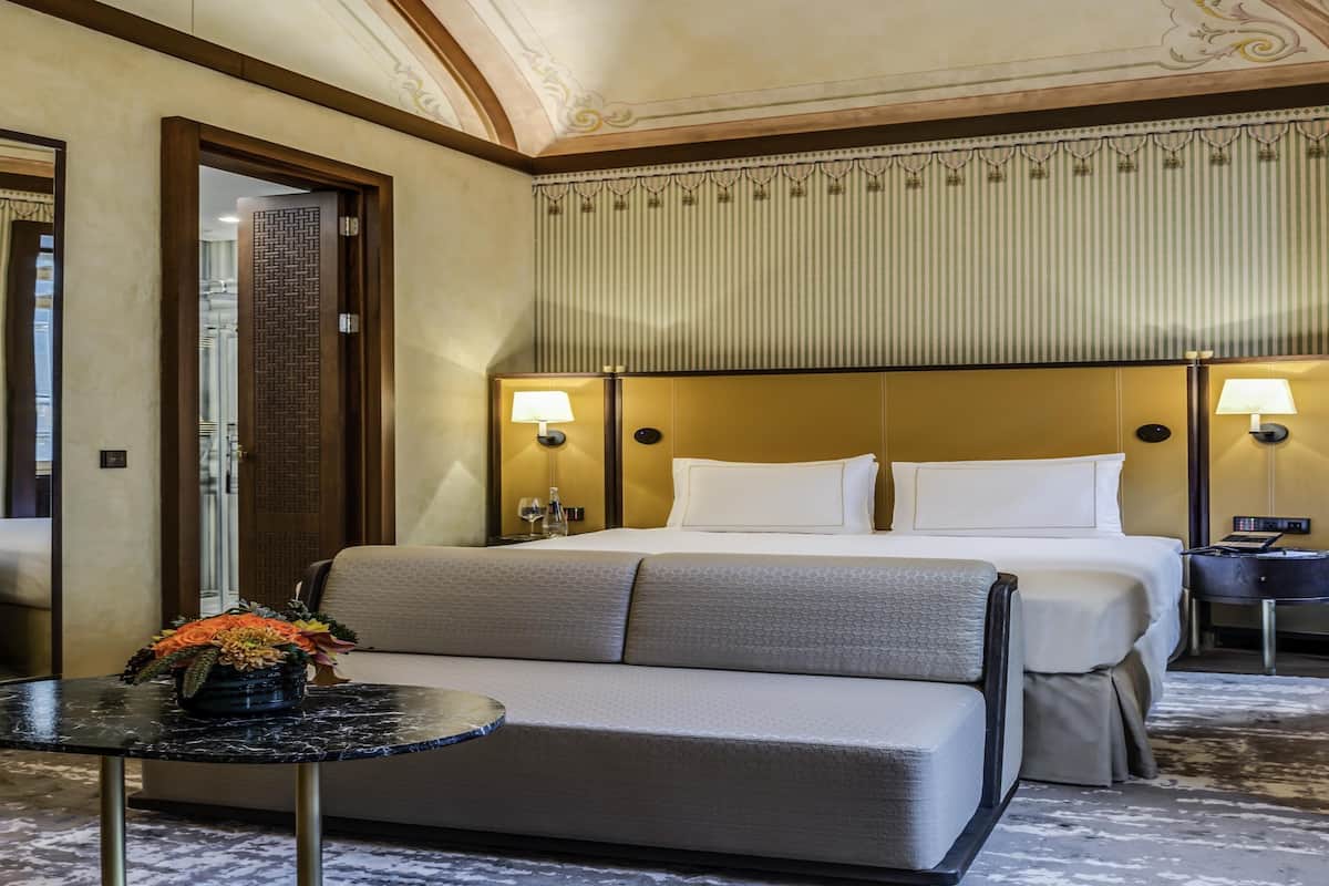 Ein Hotelzimmer mit Bett, Nachttischlampen, einem kleinen Tisch mit Blumenvase und einem Sofa.